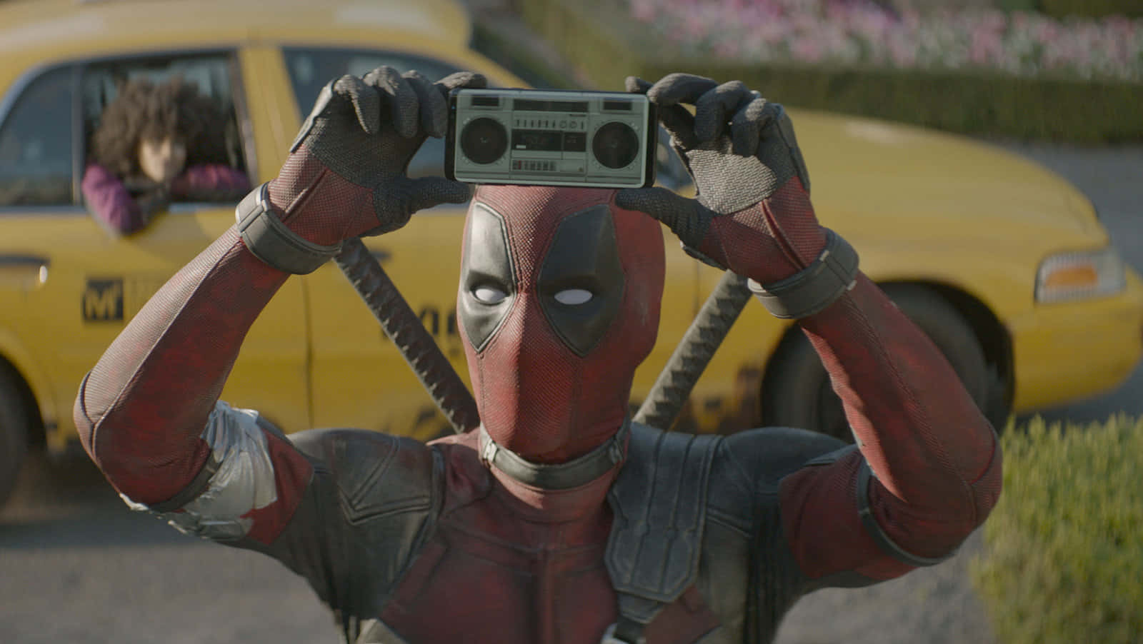 Deadpool 2 桌布 壁紙 — 死侍在黃色計程車前將一個手提音響高舉過頭，背景中可見一名乘客。