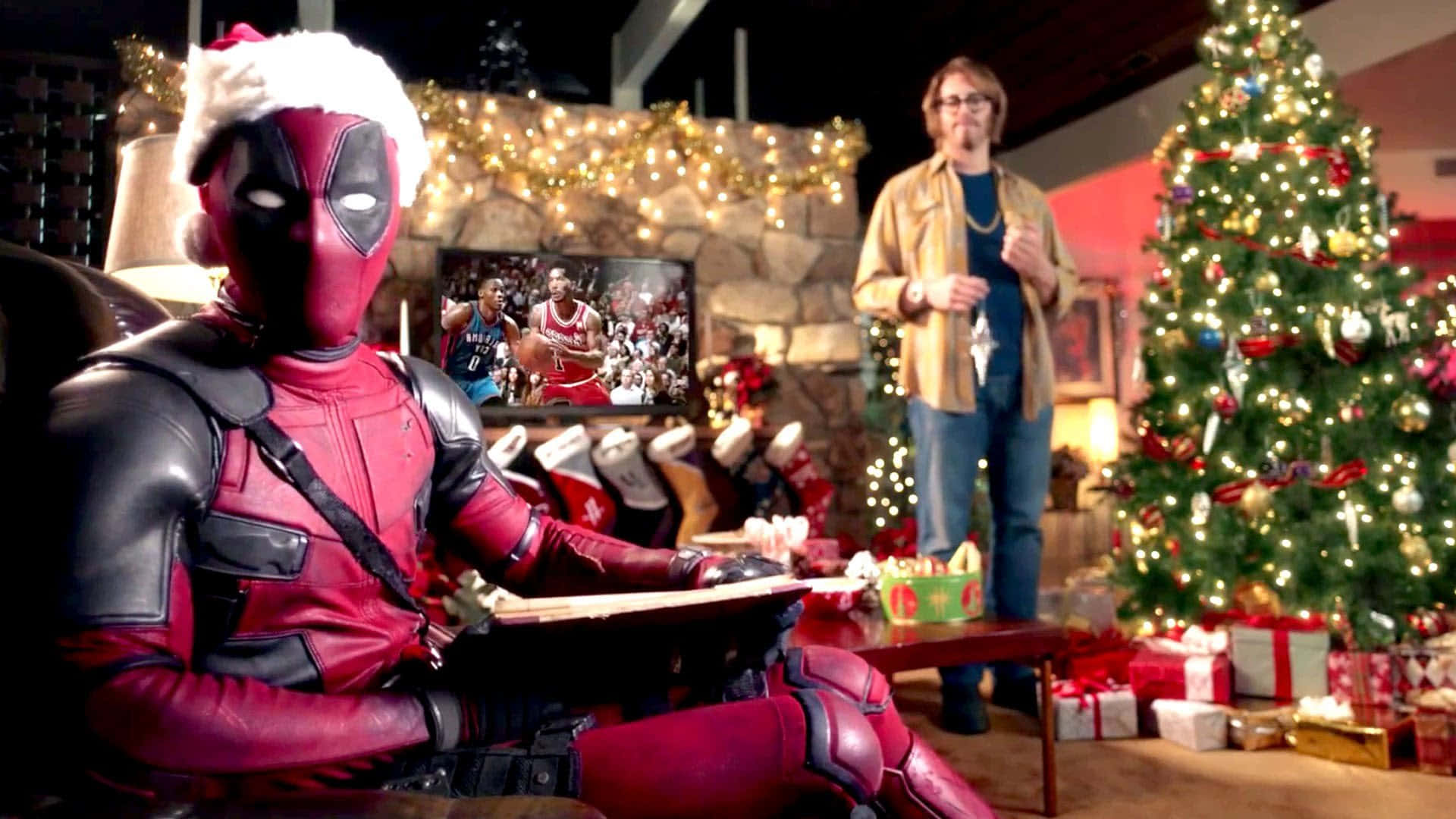 Marvel Christmas 桌布 壁紙 — 死侍戴著聖誕帽，在一個舒適的聖誕客廳裡放鬆，客廳裡有棵裝飾好的聖誕樹，還有一個男人在看電視播的籃球比賽。
