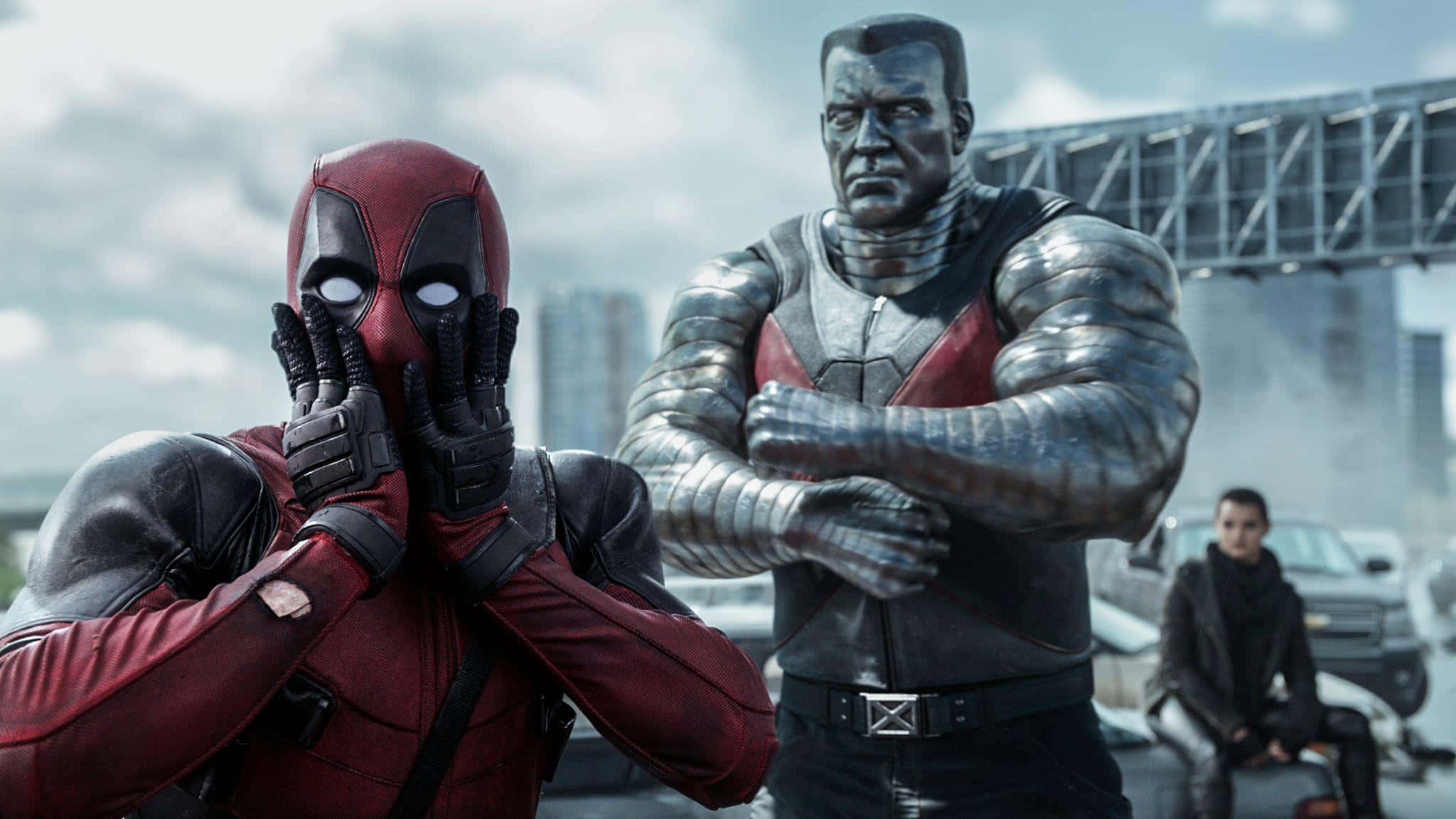 Deadpool 2 桌布 壁紙 — 死侍身穿紅色戰衣，戴著手套的雙手遮住臉部，而機堡則身穿金屬裝甲，雙臂交叉，站在城市背景前。