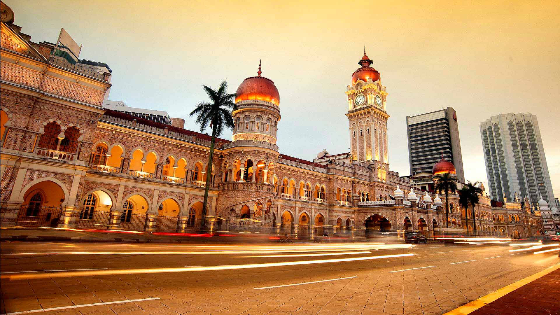 Kuala 桌布 壁紙 — 殖民時代的大型鐘樓建築在黃昏時亮起，繁忙的城市街道上車燈劃過。