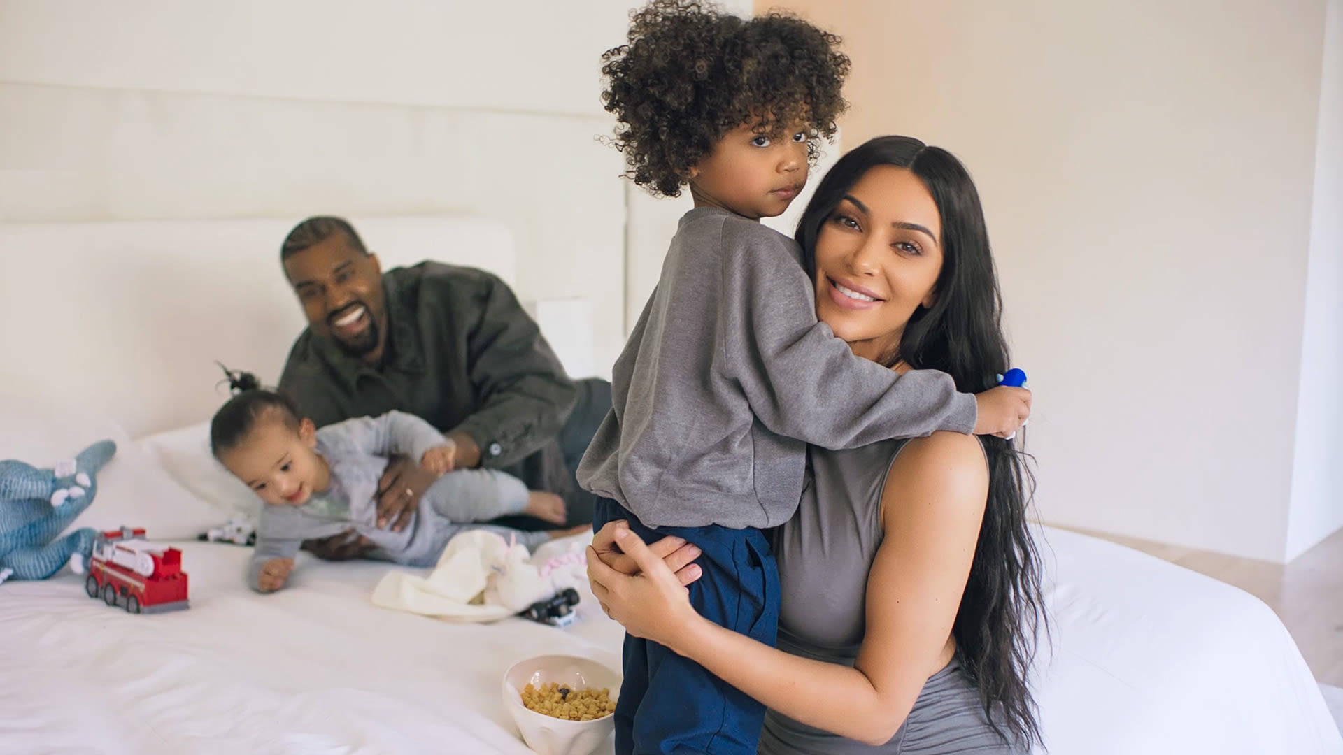Kim Kardashian 桌布 壁紙 — 母親在床上抱著孩子，父親在明亮的白色房間裡與幼兒玩耍。