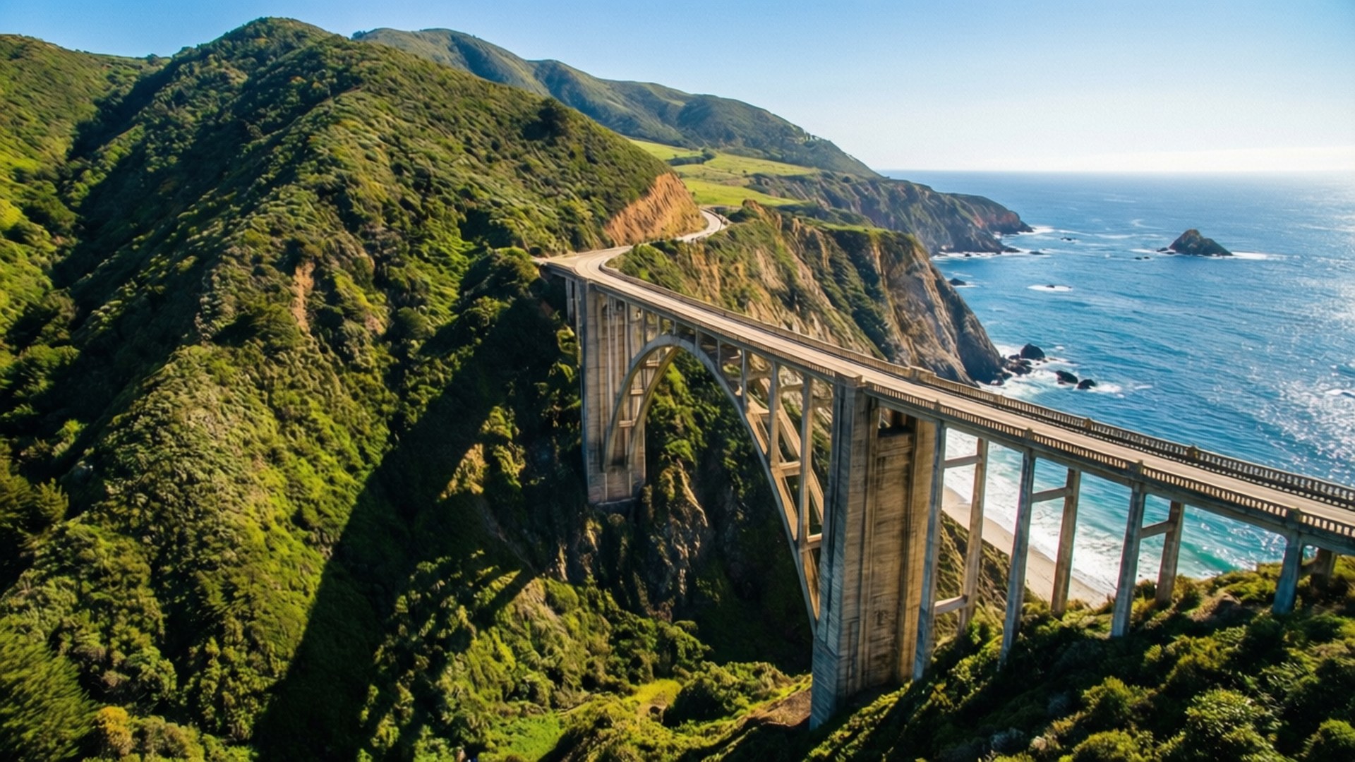 比克斯比橋全景 壁紙 — 一座雄偉的混凝土拱橋橫跨崎嶇的海岸線，下方是鬱鬱蔥蔥的綠色山丘和碧綠的海洋，上方是清澈的藍天。