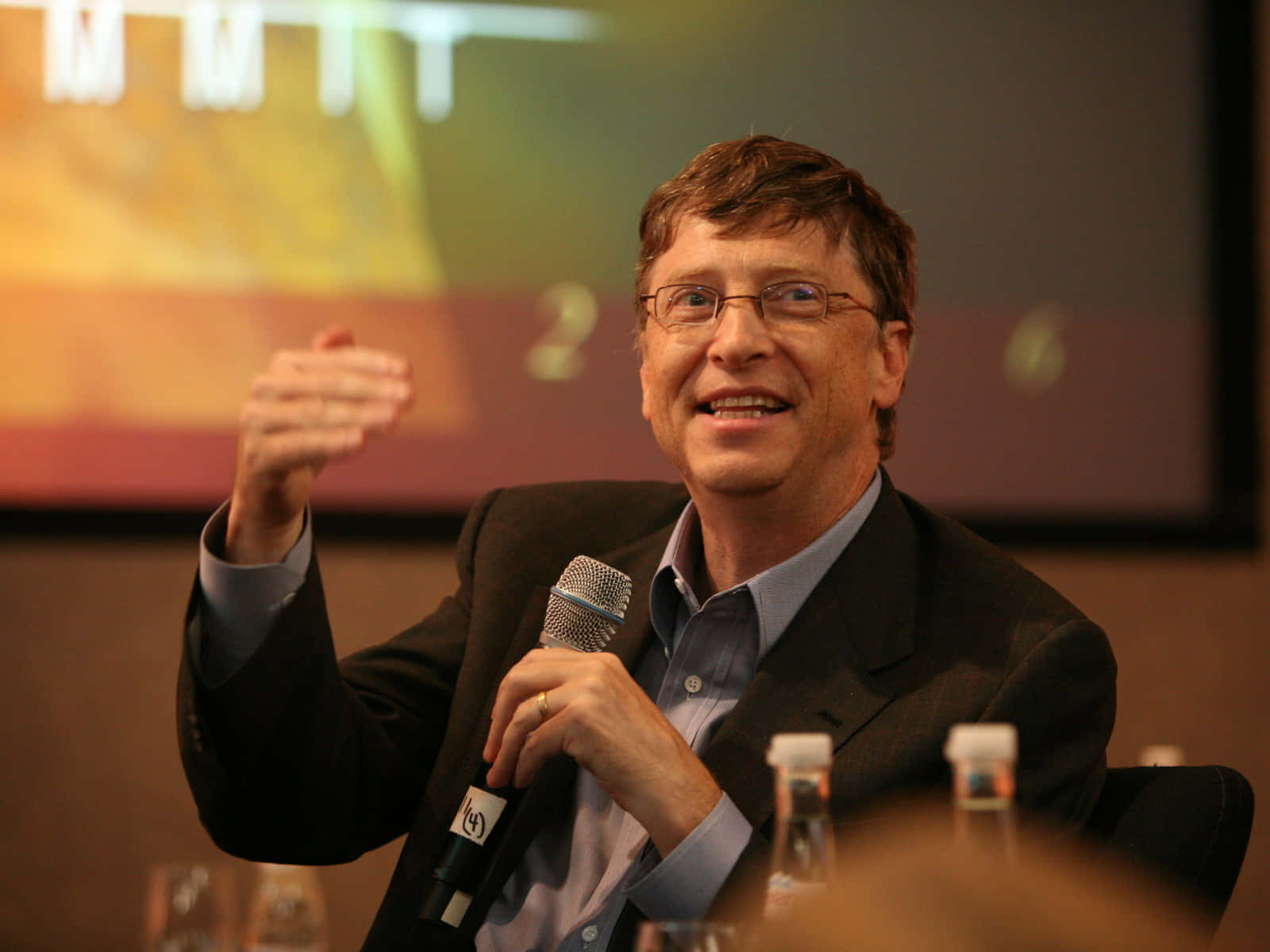 Bill Gates 桌布 壁紙 — 比爾·蓋茲在會議上對著麥克風溫暖地微笑說話，身後螢幕上可見「MIT」字樣。