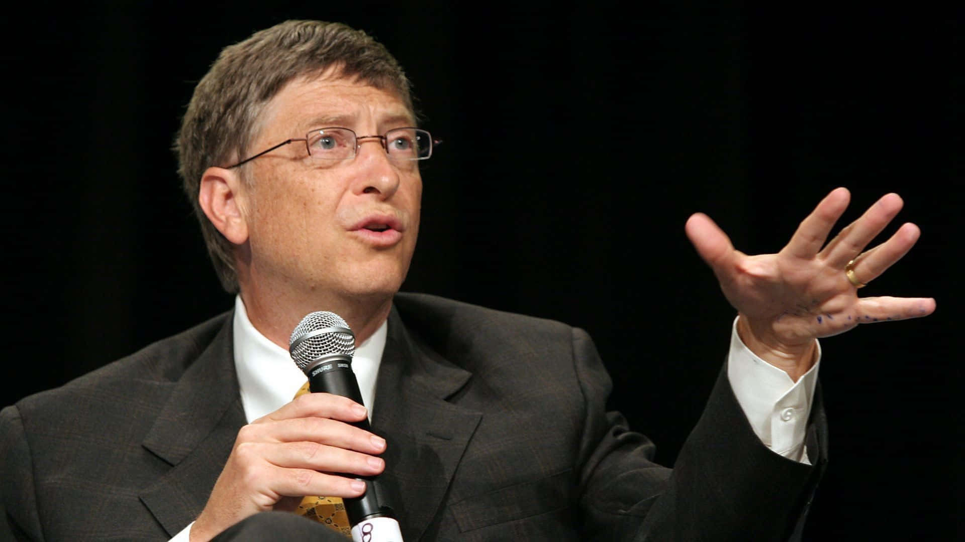 Bill Gates 桌布 壁紙 — 比爾·蓋茲在深色背景前用左手做手勢對著麥克風講話。