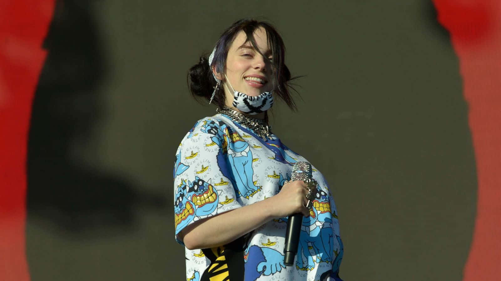 Billie Eilish Laptop 桌布 壁紙 — 比莉·艾利許在舞台上燦爛微笑，身穿藍色卡通圖案襯衫，手持麥克風。