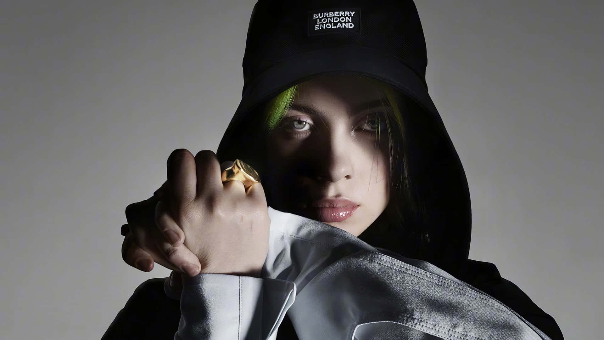 Billie Eilish Laptop 桌布 壁紙 — 比莉·艾利許戴著黑色 Burberry 漁夫帽，露出螢光綠色頭髮，在純灰色背景前凝視。