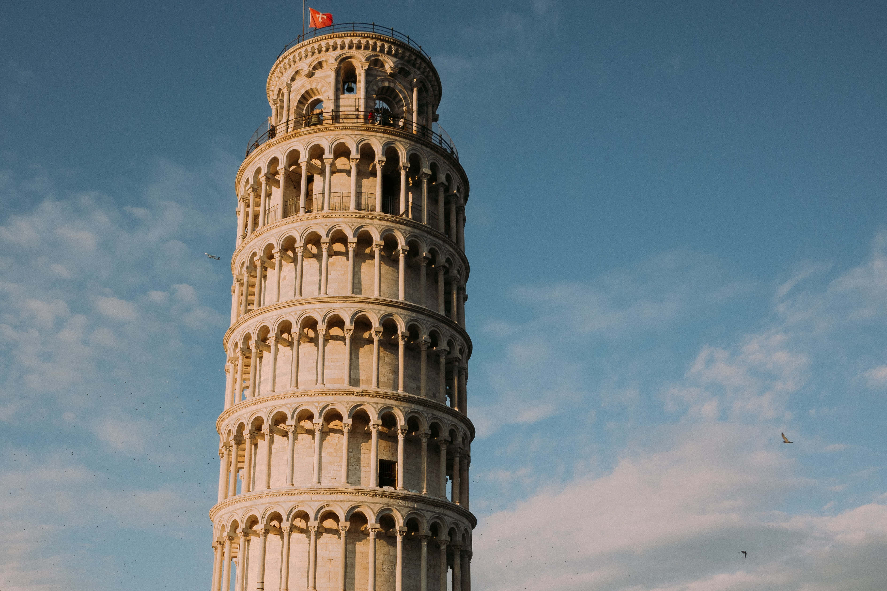 Italy Aesthetic 桌布 壁紙 — 比薩斜塔在明亮的藍天下高高聳立，天空中有縷縷白雲，塔頂飄揚著一面紅旗。