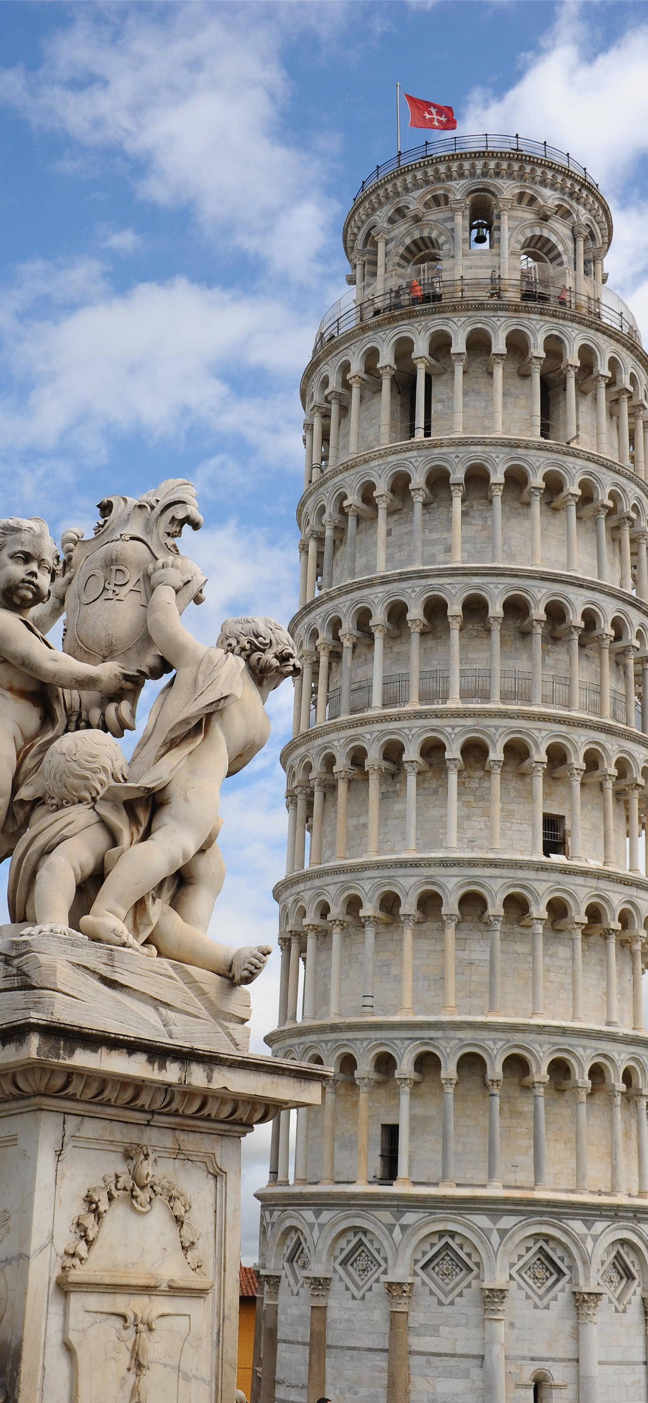 Tuscany 桌布 壁紙 — 比薩斜塔高聳在明亮的藍天和散落的雲朵之間，其歷史悠久的石造建築和頂部的旗幟與前景中精緻的大理石雕像形成對比。