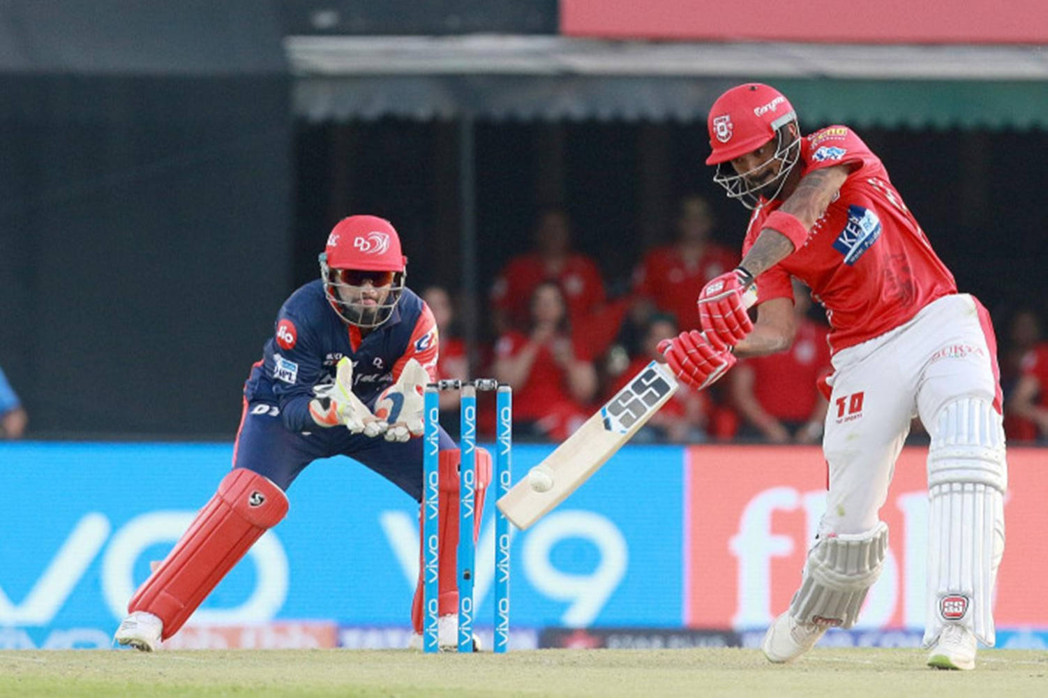 kl rahul 桌布 壁紙 — 比賽中，一名身穿紅色的板球擊球手揮動球棒擊球，而一名身穿藍色的守門員蹲在柱門後面。