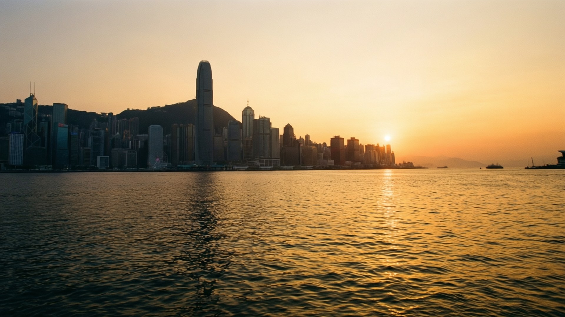 水面上的金色日出 壁紙 — 金色夕陽照耀香港天際線，倒映在平靜的水面上，遠處有船隻。