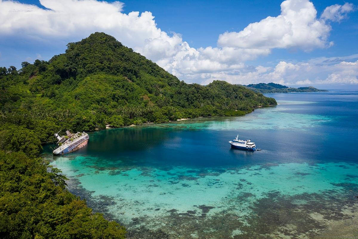 山景 桌布 壁紙 — 沉沒的遊輪擱淺在鬱鬱蔥蔥的綠色島嶼海岸旁的鳥瞰圖，旁邊是碧綠的海水，附近有一艘小船。