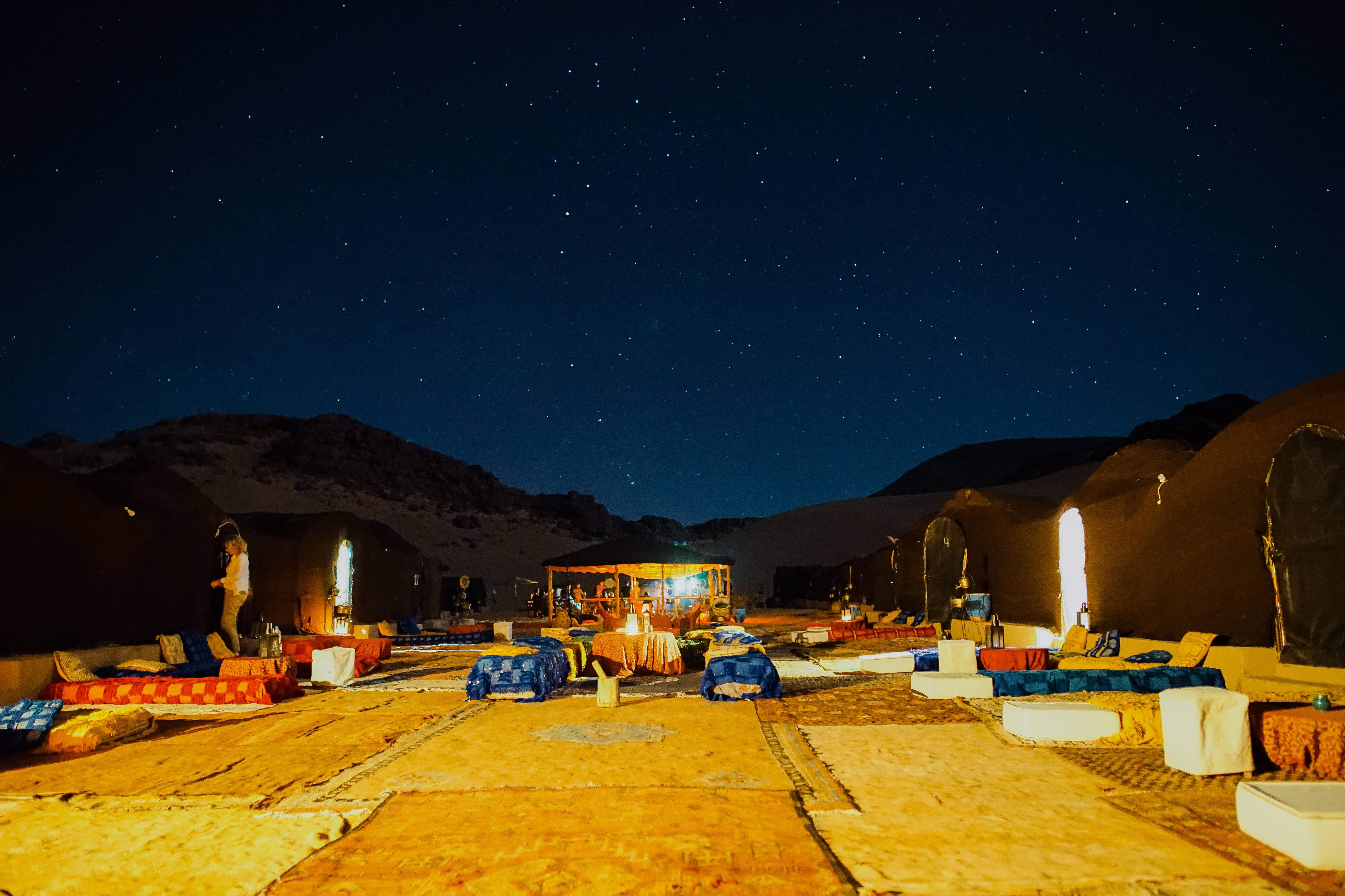 Sahara 桌布 壁紙 — 沙漠營地在繁星點點的夜空下溫暖地發光，背景有山脈。