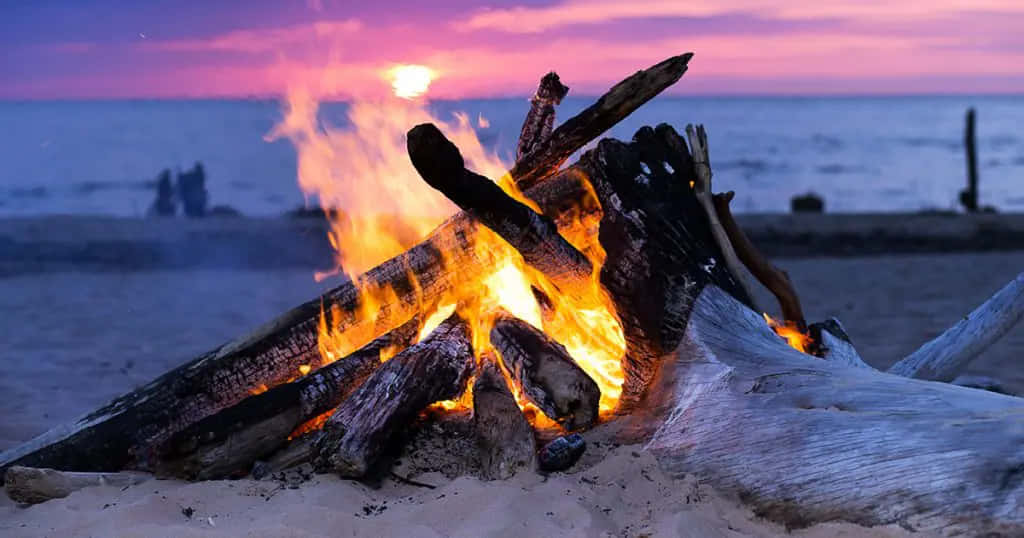 Bonfire 桌布 壁紙 — 沙灘上，營火在日落時分熊熊燃燒，在紫色和橙色的天空映襯下散發出溫暖的光芒。