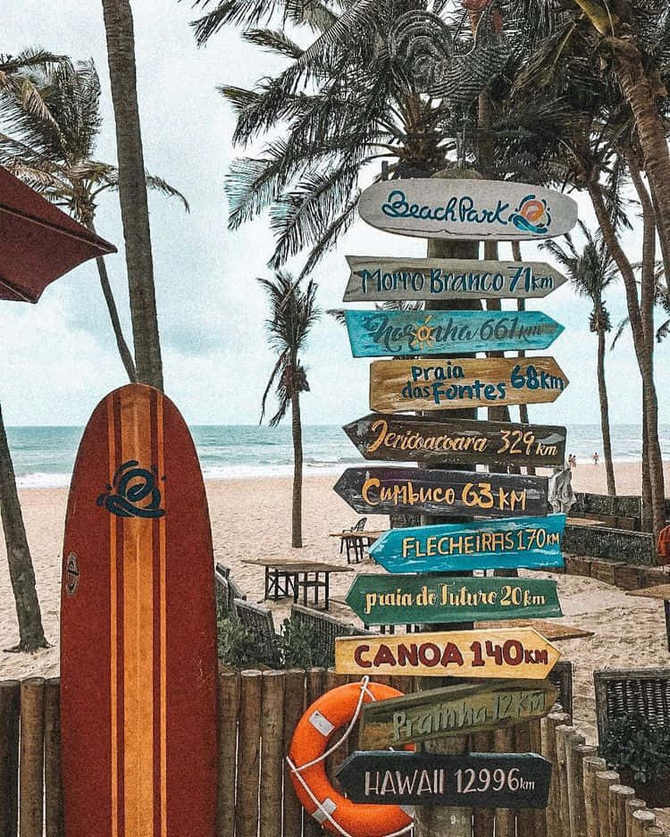 surfing 桌布 壁紙 — 沙灘上一個色彩繽紛的木製路標，指向遙遠的海岸城鎮和棕櫚樹下的夏威夷，旁邊靠著一塊衝浪板。