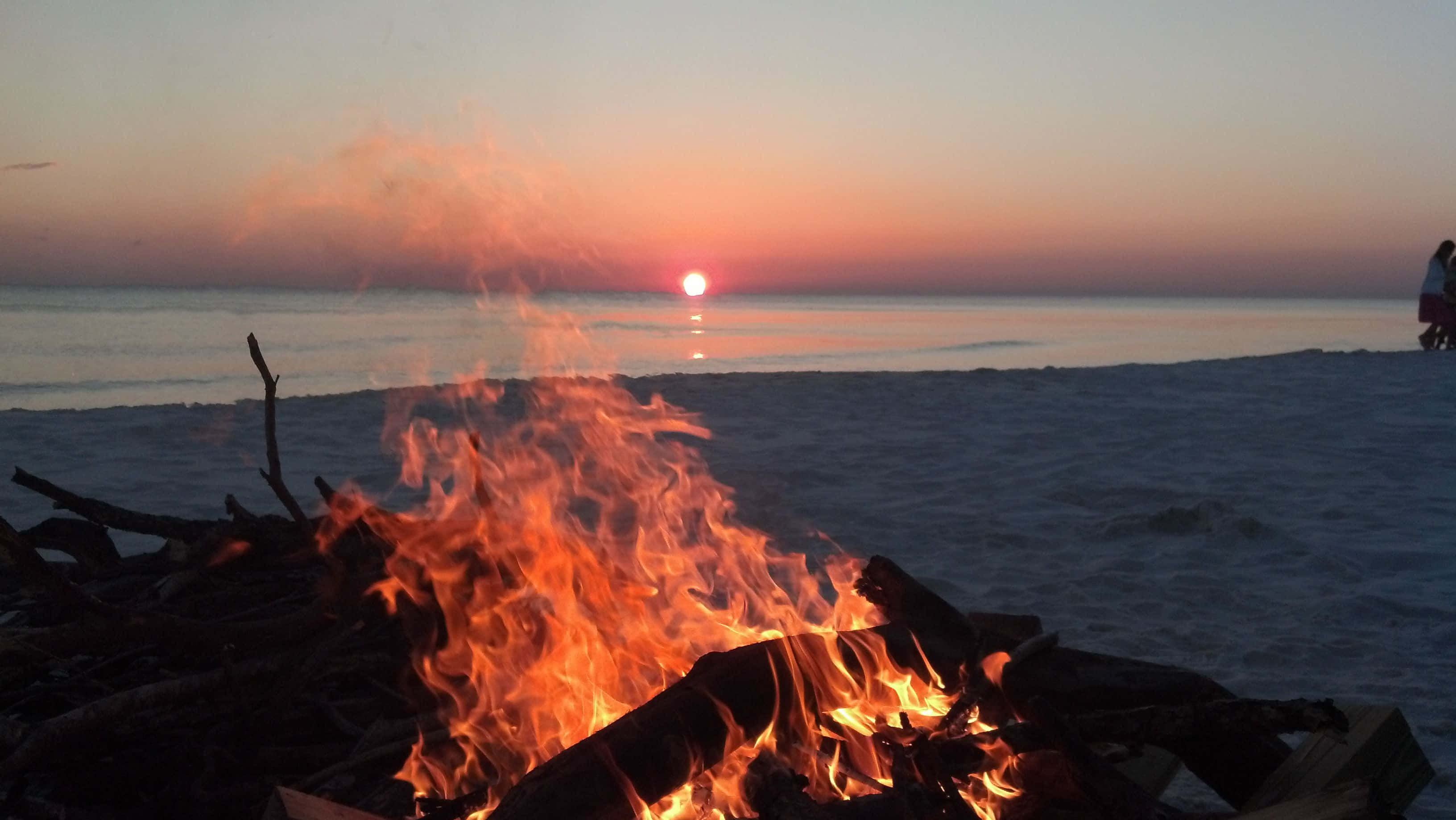 Beach Bonfire 桌布 壁紙 — 沙灘上熊熊燃燒的營火在日落天空下舞動，兩個人影在海岸上形成剪影。
