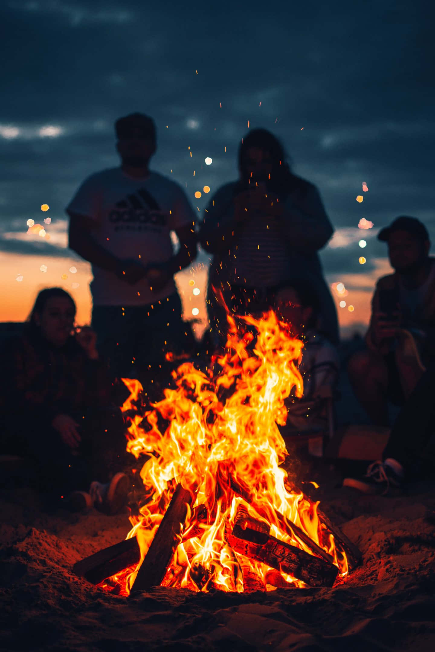 Bonfire 桌布 壁紙 — 沙灘上營火劈啪作響，朋友們聚集在暮色天空下，周圍有發光的餘燼和遠處的燈光。