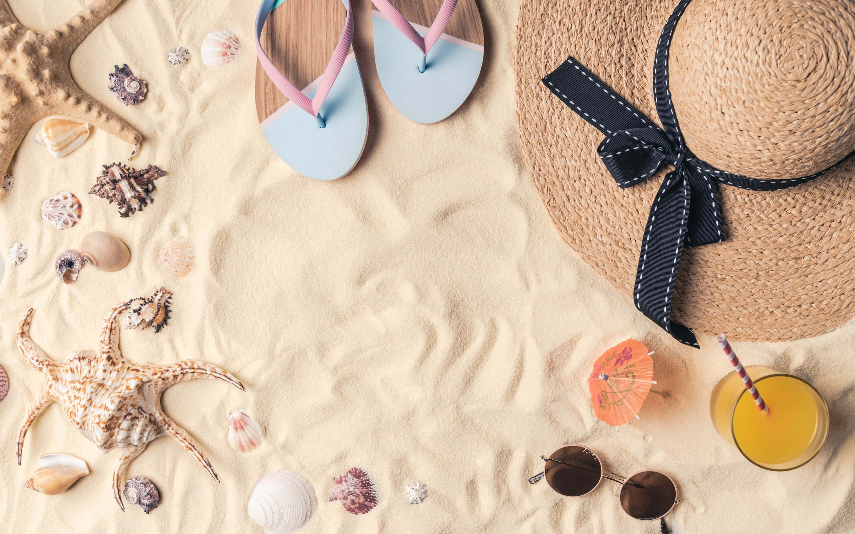 Beach Hat 桌布 壁紙 — 沙灘必需品平鋪擺放，包括人字拖、草帽、太陽眼鏡和熱帶飲品，置於柔軟沙灘上。