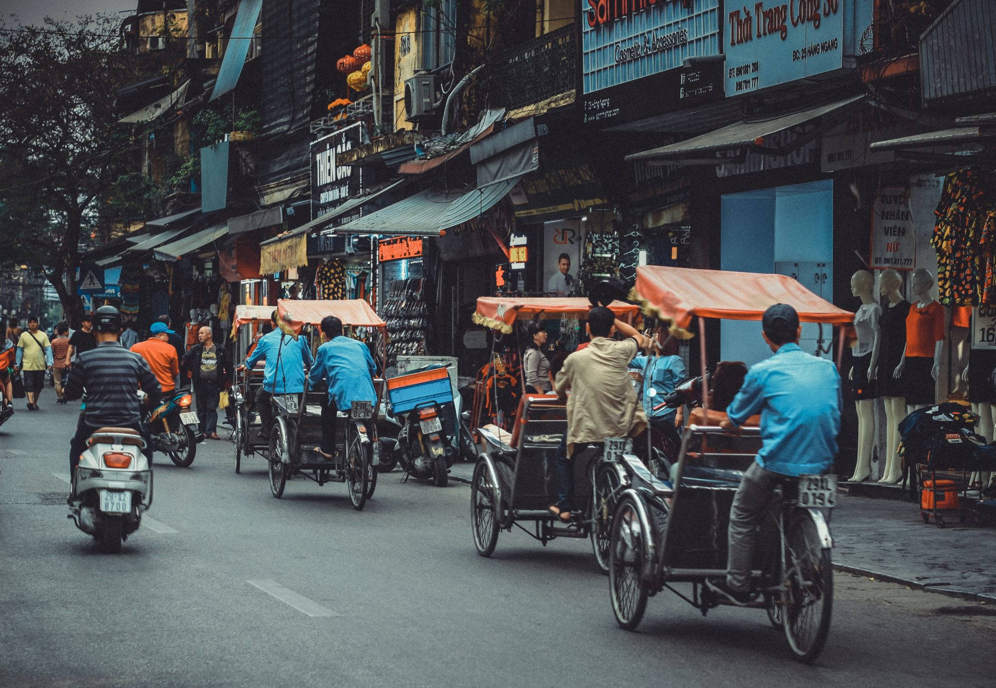 河內 桌布 壁紙 — 河內繁忙的街景，摩托車和人力車在狹窄的小巷中穿梭，兩旁商店林立，擺放著人體模型。