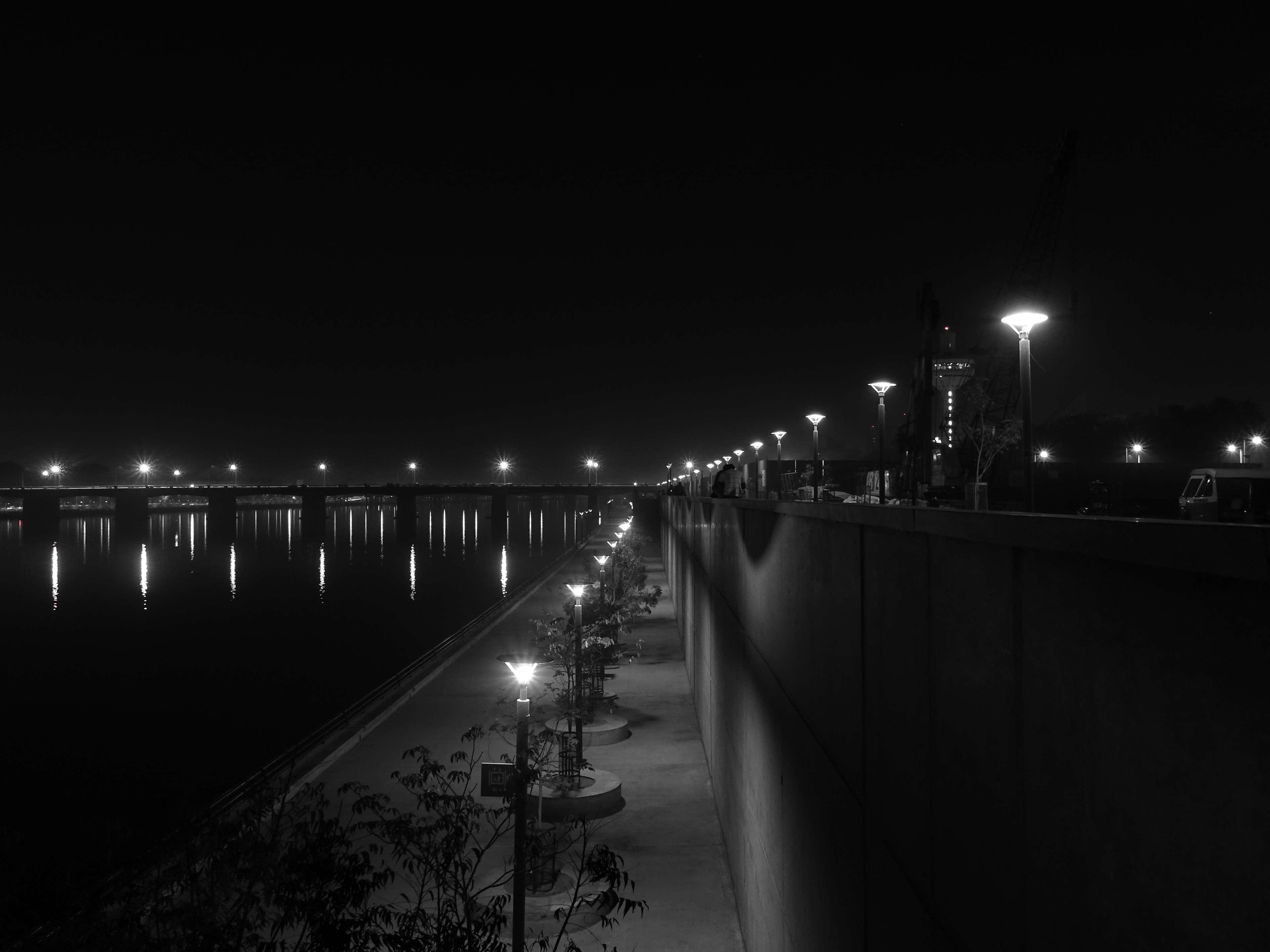 Black And White City 桌布 壁紙 — 河岸步道夜景黑白相片，燈光璀璨的街燈倒映在平靜水面上。