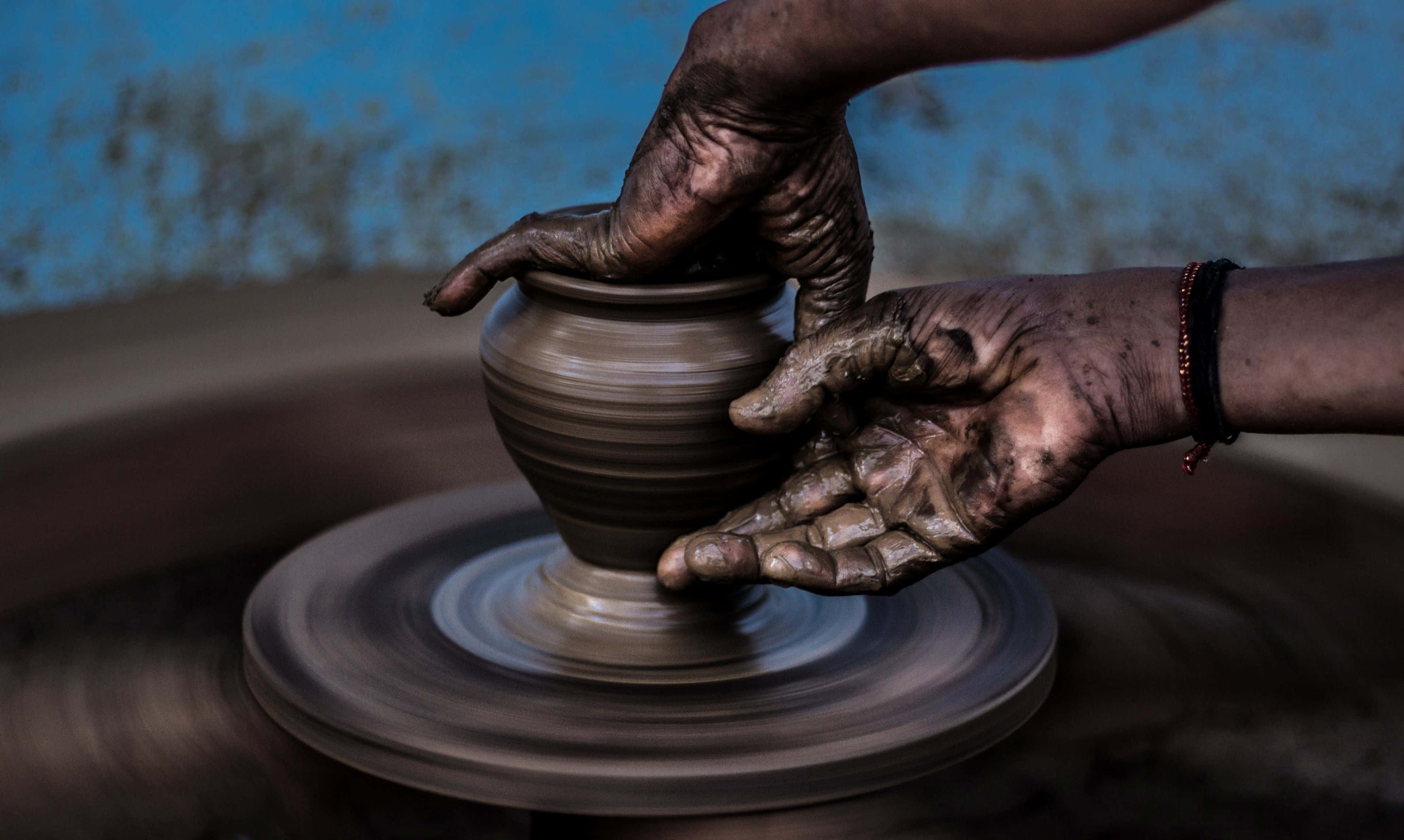 Ceramics 桌布 壁紙 — 沾滿泥巴的雙手在紡車上塑造陶罐，背景是帶紋理的藍色牆壁。