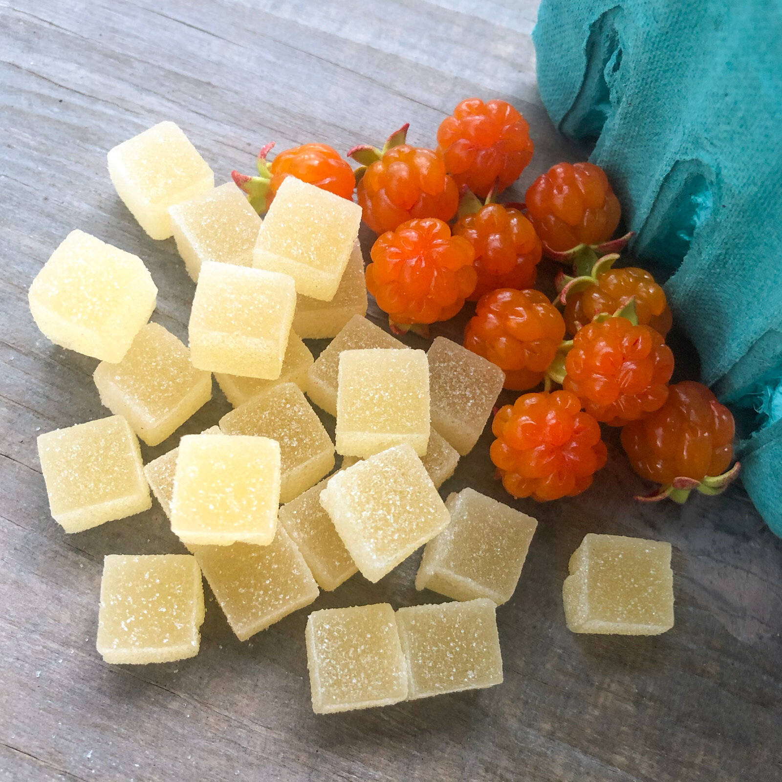 Berry 桌布 壁紙 — 沾滿糖的黃色糖果方塊和鮮豔的橙色覆盆子放在經風化的木頭上，旁邊有一塊青綠色的布。