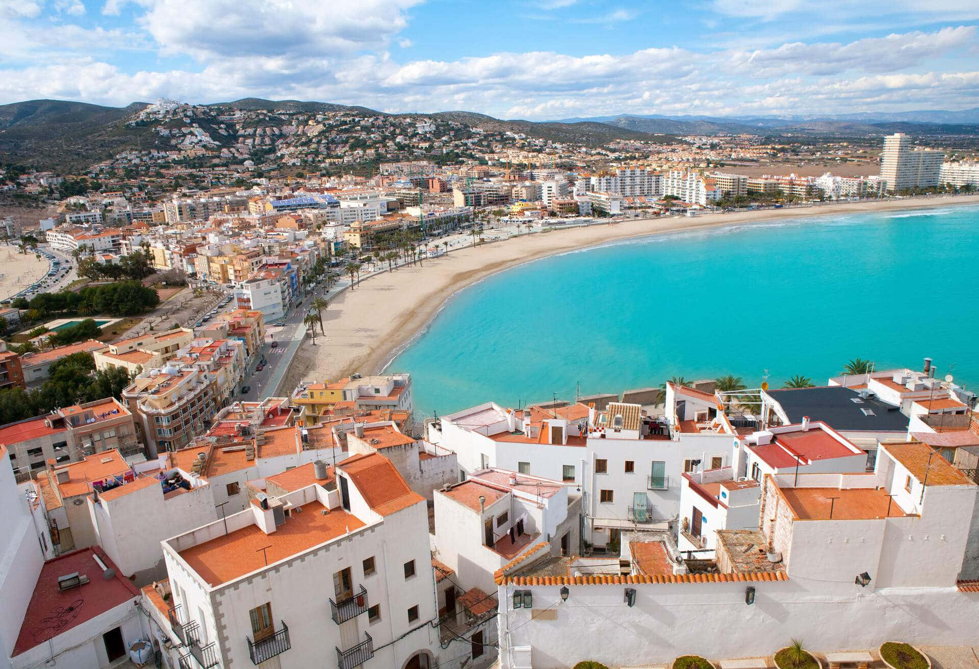 Spanish Beach 桌布 壁紙 — 沿海城鎮的鳥瞰圖，白色建築和陶土屋頂環抱著碧綠的海灣，天空部分多雲。