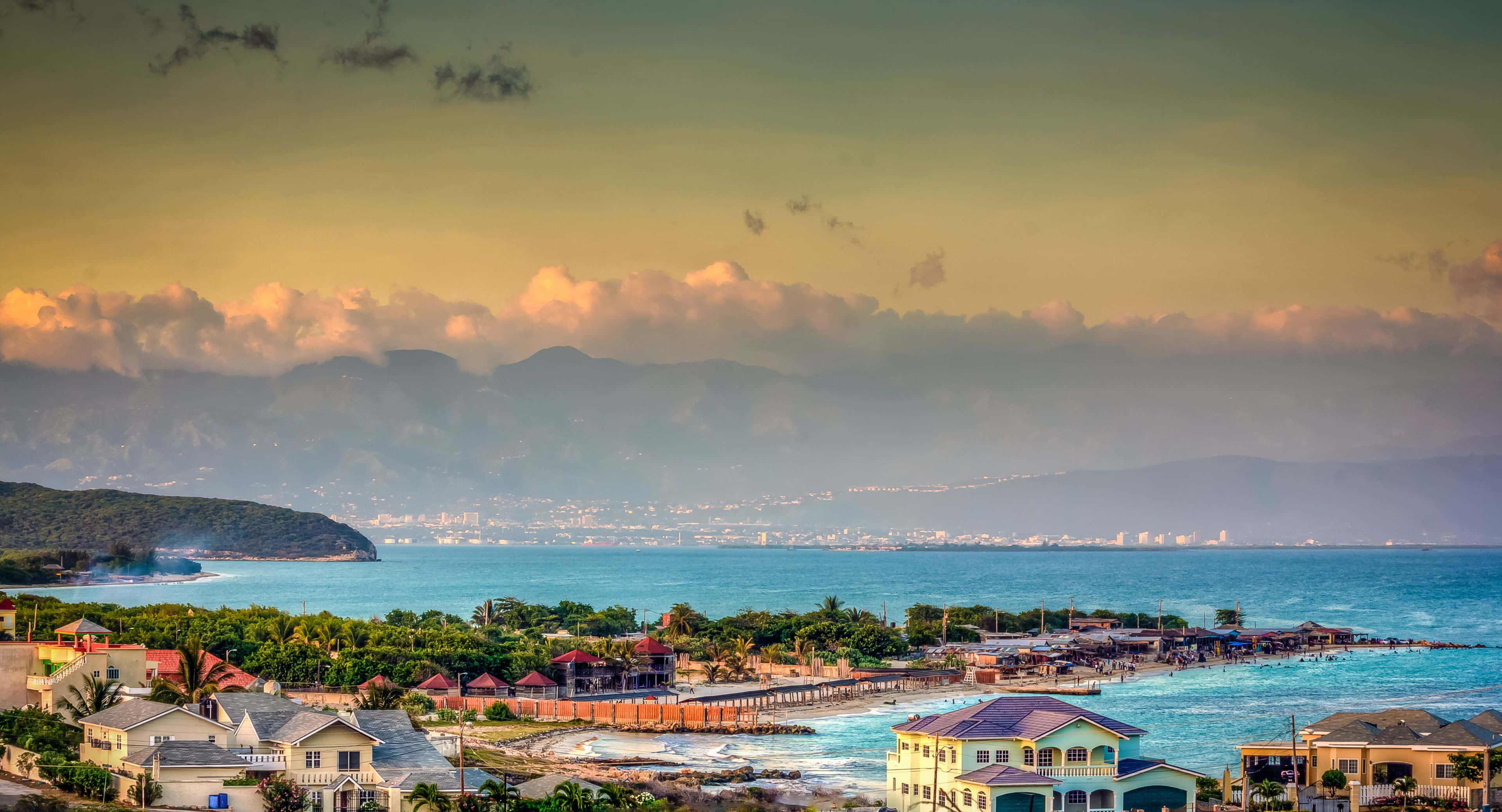 3840x2076 桌布 壁紙 — 沿海小鎮，粉彩房屋和棕櫚樹依偎在碧綠的海水旁，遠方山巒在日落時分。