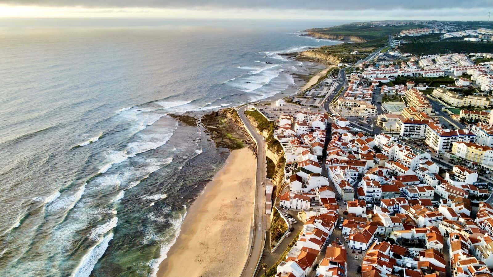 Portuguese Beach 桌布 壁紙 — 沿海小鎮的鳥瞰圖，白色建築和紅色屋頂依偎在沙灘和拍岸的海浪旁。