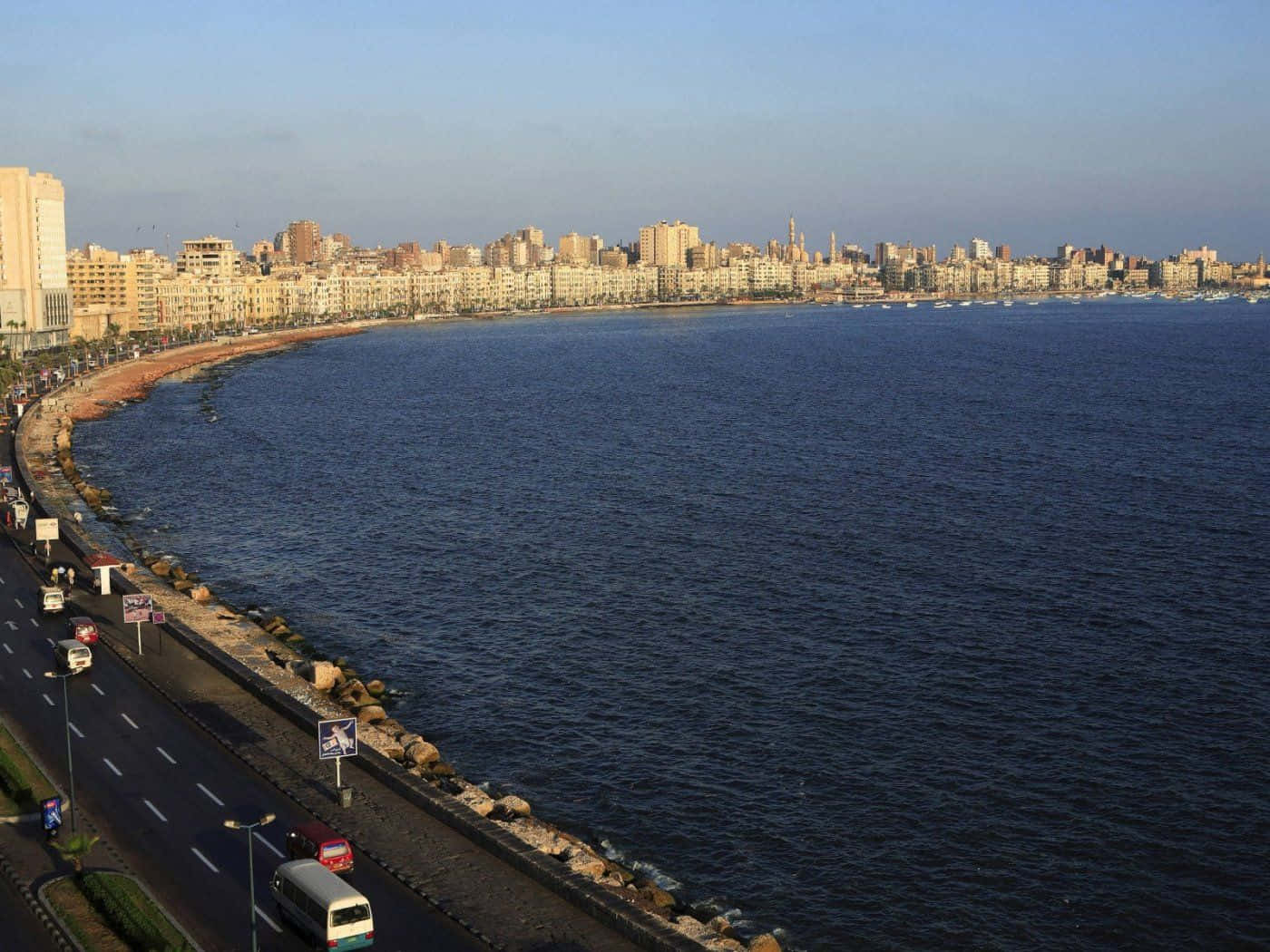 Alexandria 桌布 壁紙 — 沿著彎曲海岸線的繁華城市景觀，寬闊的海岸景色，繁忙的道路和深藍色的海水在晴朗的天空下。