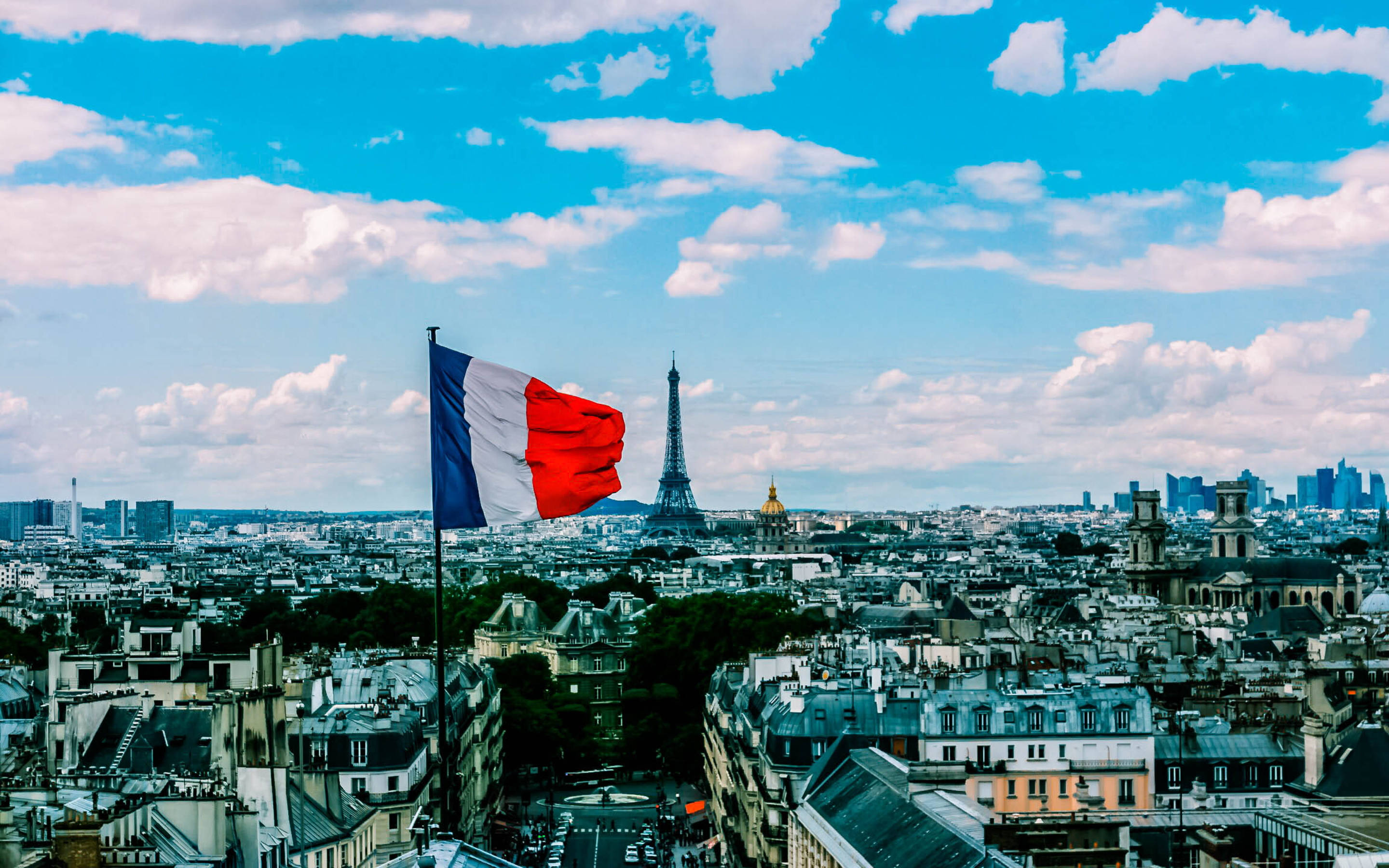 France Flag 桌布 壁紙 — 法國國旗在巴黎天際線上自豪地飄揚，艾菲爾鐵塔在點綴著蓬鬆雲朵的明亮藍天下高高聳立。