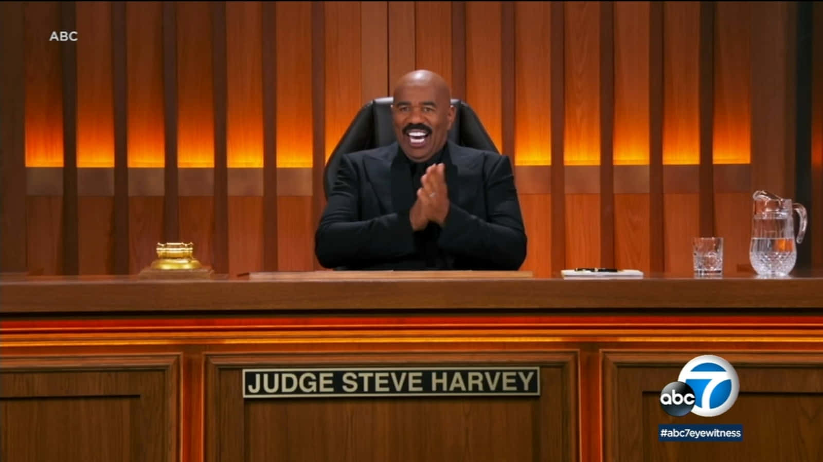 steve harvey 桌布 壁紙 — 法官Steve Harvey坐在木製長凳後面，在溫暖照明的法庭環境中咧嘴笑著拍手。
