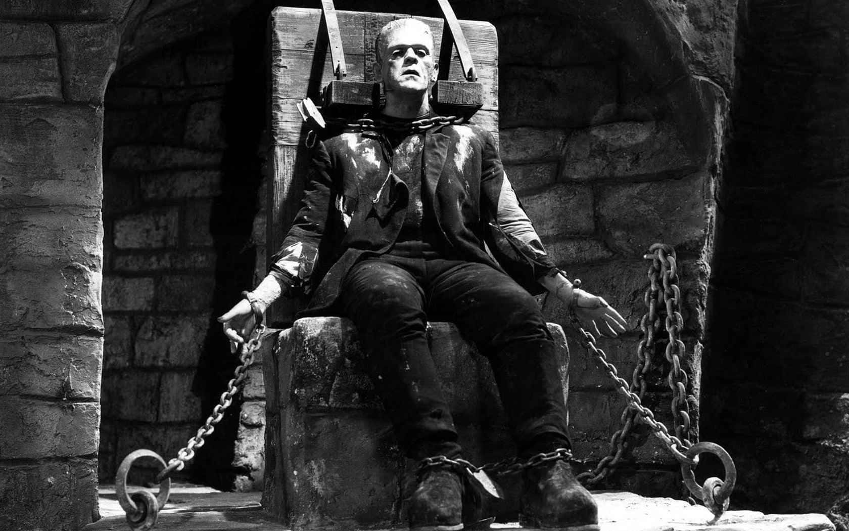 Frankenstein 桌布 壁紙 — 法蘭克斯坦的怪物被鎖鏈鎖在石頭地牢中，脖子上戴著沉重的木製頸圈，手腕和腳踝也纏著鎖鏈。