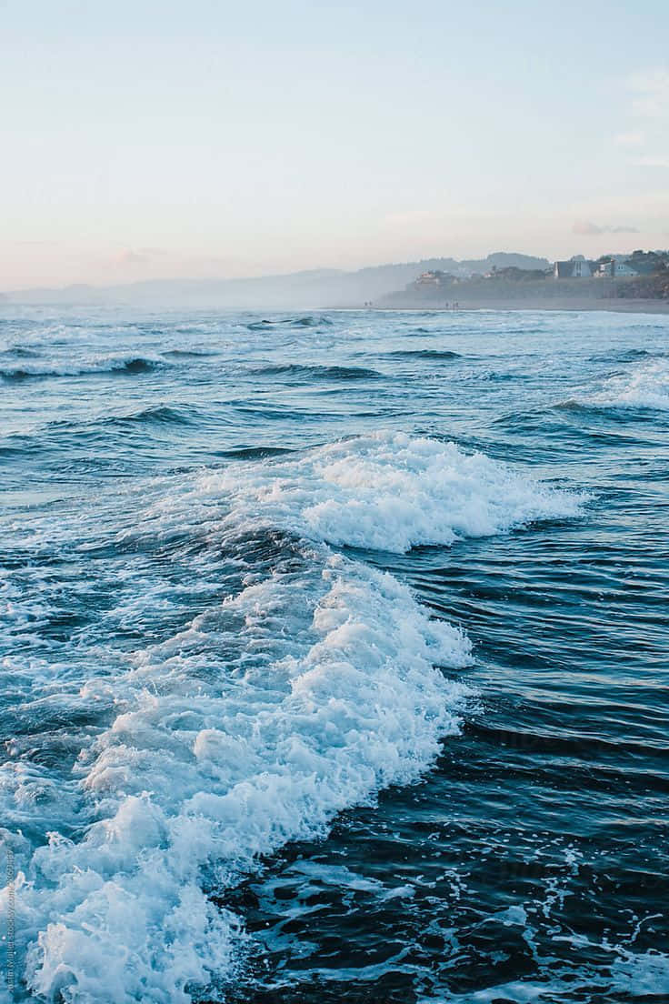 Sea Aesthetic 桌布 壁紙 — 泡沫海浪在柔和粉彩天空下，伴隨著遠處海岸山丘和建築物，向岸邊翻滾。