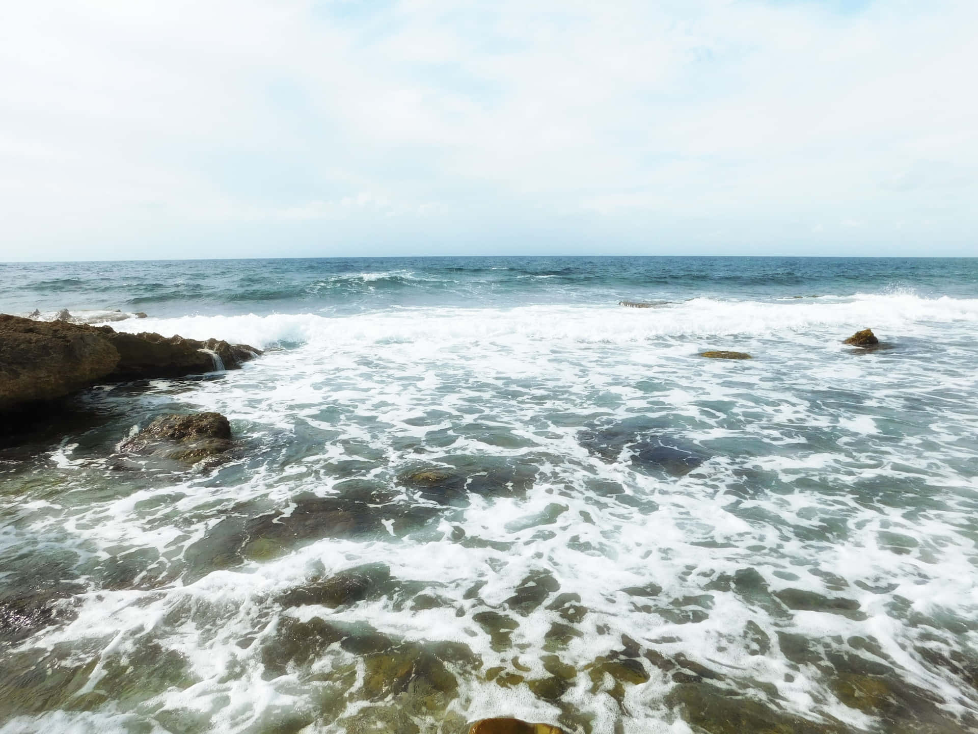 Sea 壁紙 — 泡沫海浪拍打深色岩石，天空淡藍，雲朵散落。
