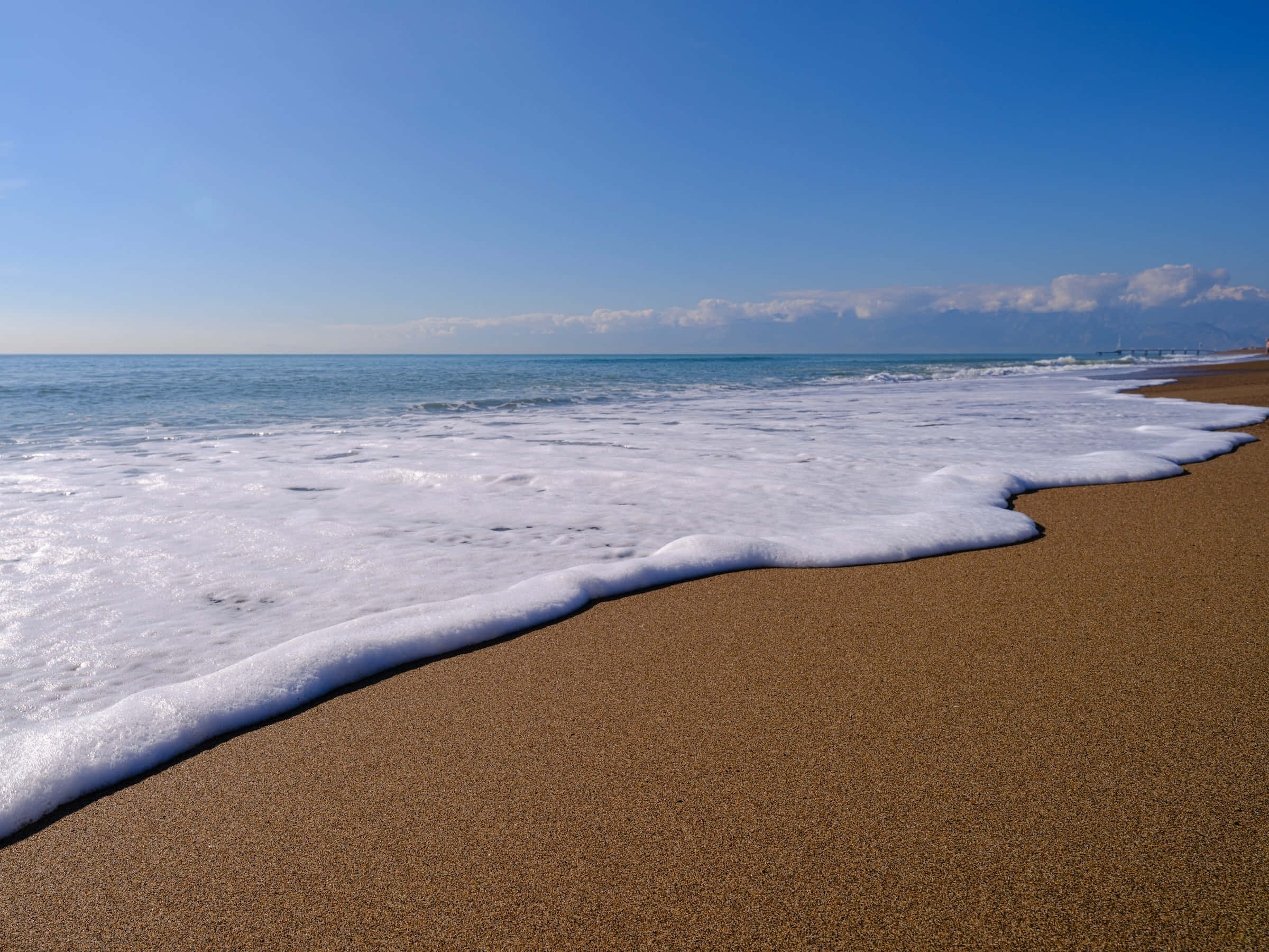 Beach Desktop 桌布 壁紙 — 泡沫狀的海浪在晴朗藍天和遠處雲朵下，輕輕拍打陽光照耀的金色沙灘。