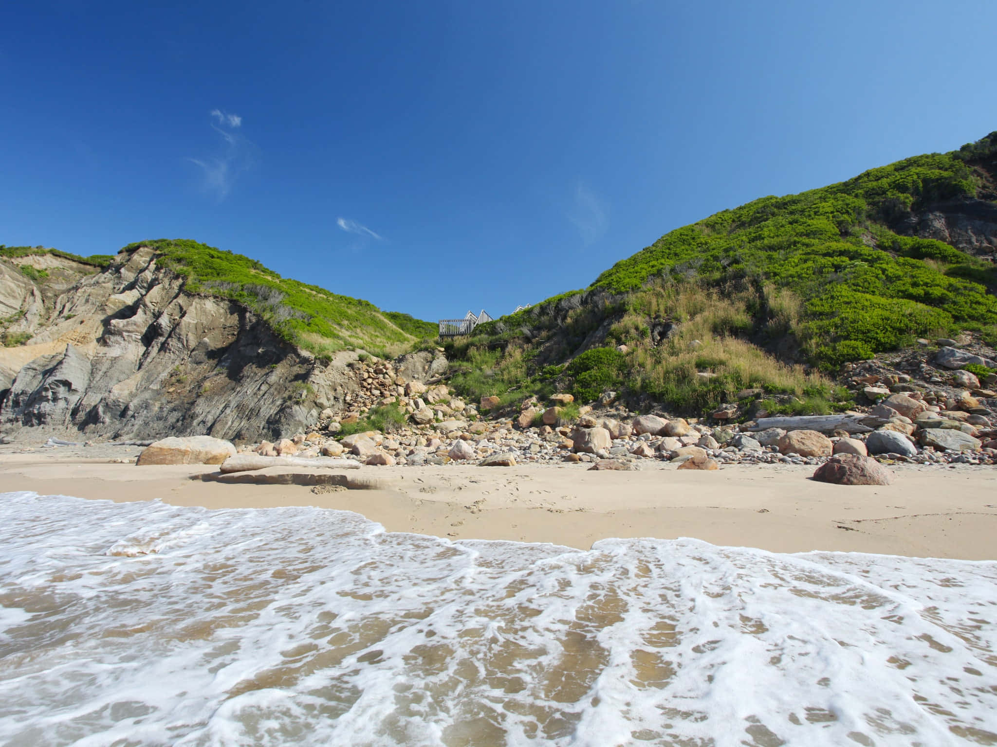 風景 桌布 壁紙 — 泡沫般的海浪拍打著沙灘，沙灘兩旁是岩石峭壁和翠綠的山丘，頭頂是清澈的藍天。