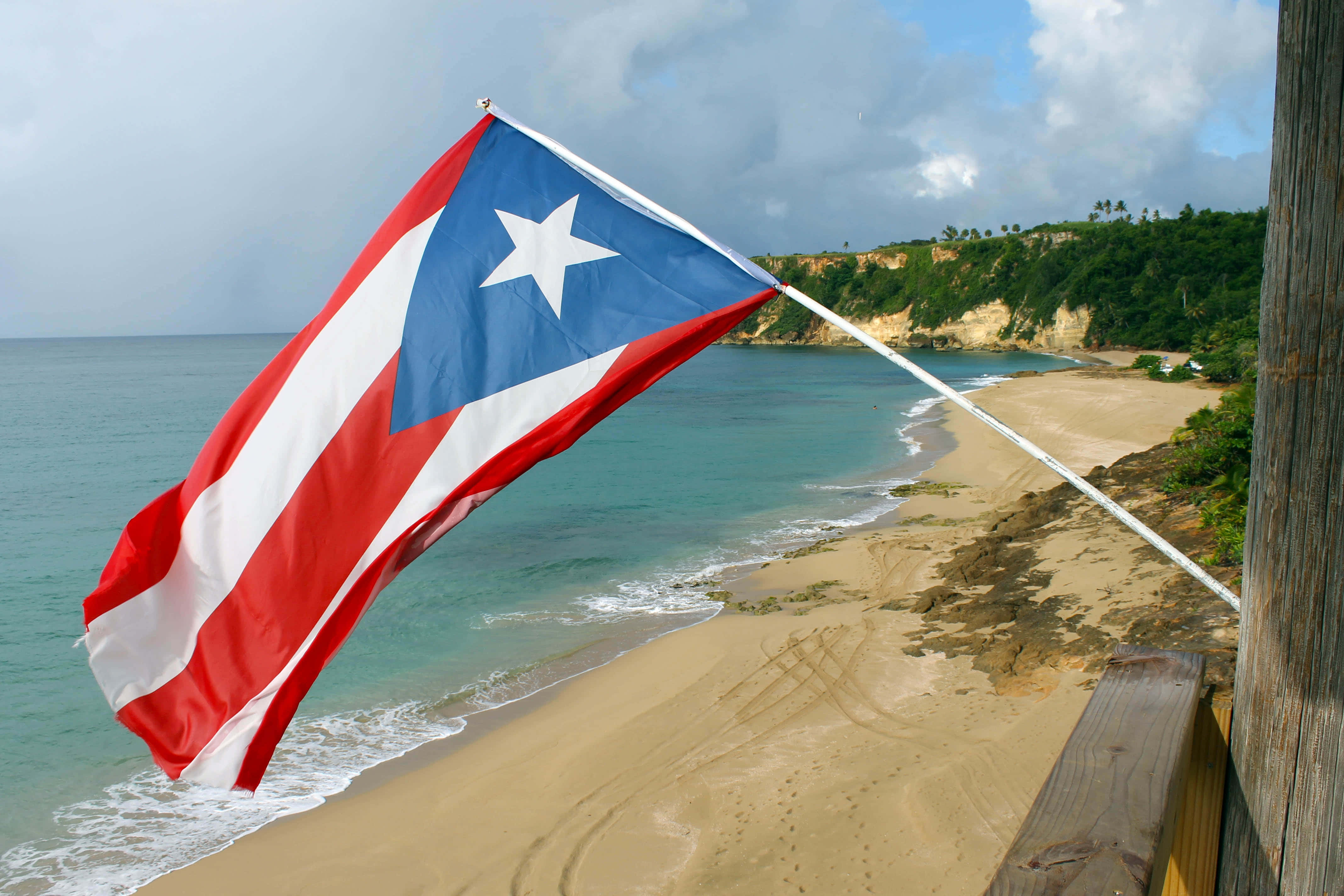 Puerto Rican 桌布 壁紙 — 波多黎各旗幟在陰天下，以藍綠色海洋和沙灘為背景，勇敢地飄揚。