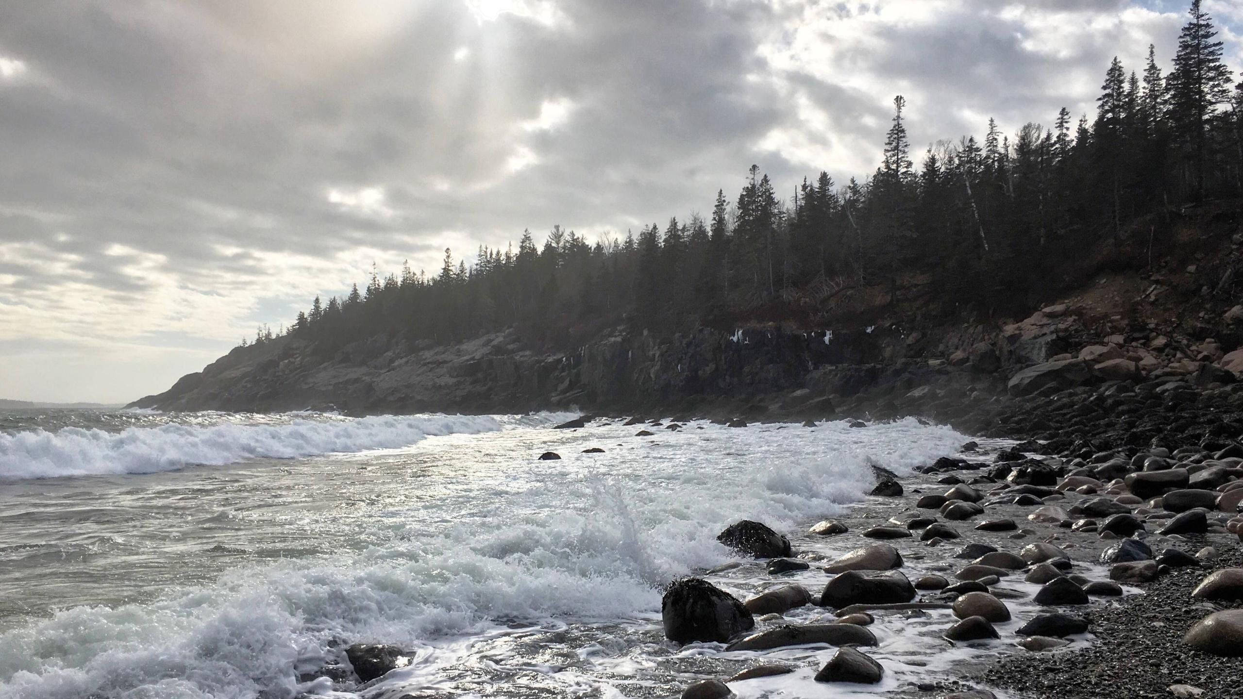 rocky 桌布 壁紙 — 波浪拍打岩石海岸，天空多雲，陽光穿過樹林覆蓋的懸崖上方。