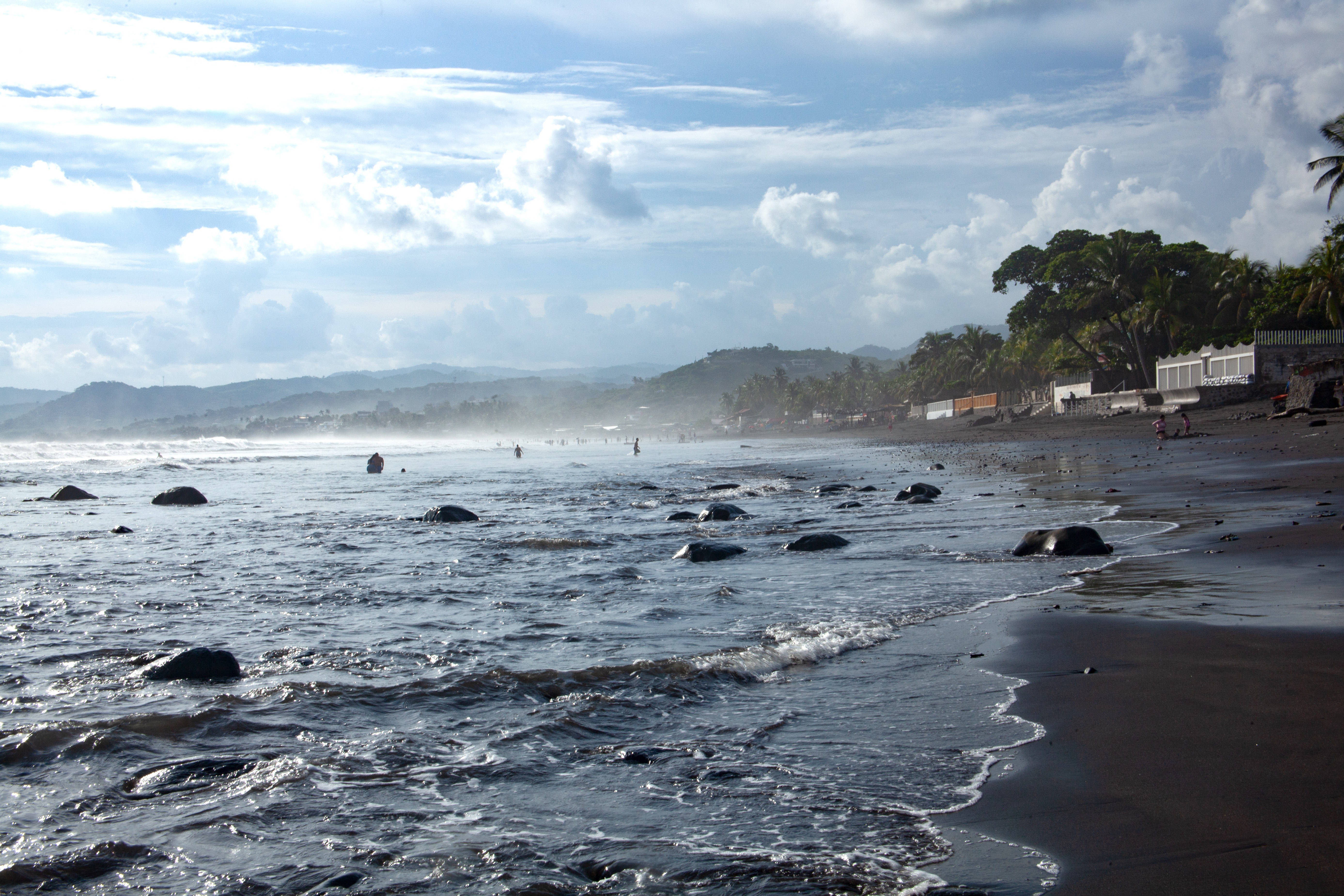 El Salvador 桌布 壁紙 — 波浪拍打著一個點綴著岩石的深色火山沙灘，天空明亮多雲，遠處有山丘和棕櫚樹。
