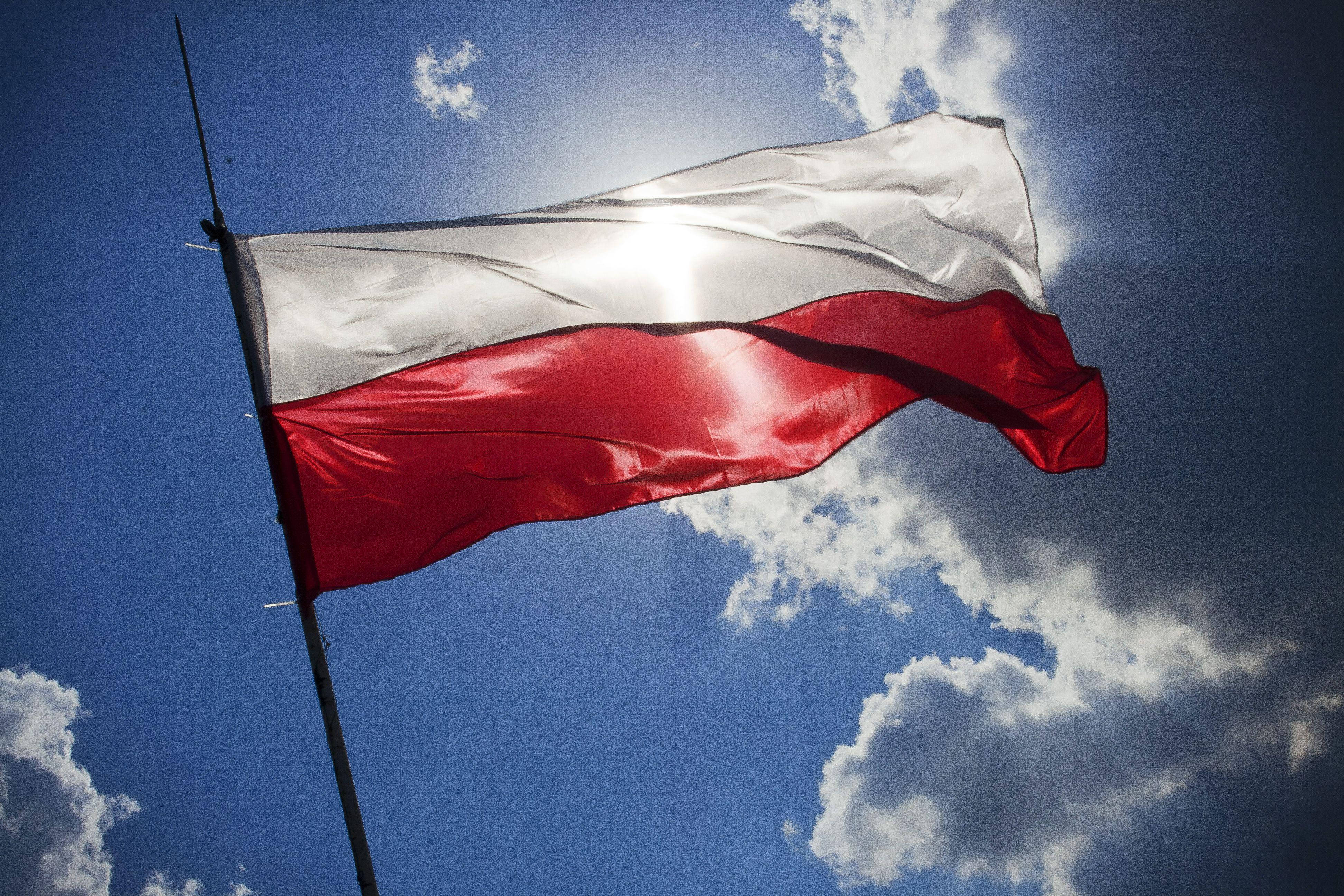 Poland Flag 桌布 壁紙 — 波蘭國旗帶有紅色和白色條紋在風中飄揚，背景是明亮的藍天，有散落的雲朵和太陽光暈。