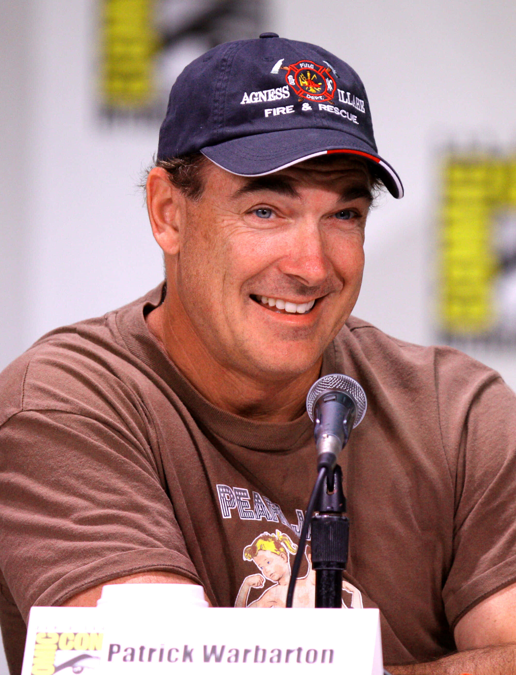 Patrick Warburton 桌布 壁紙 — 派翠克·沃伯頓在Comic-Con座談會上戴著消防救援帽和棕色T恤，溫暖地微笑。