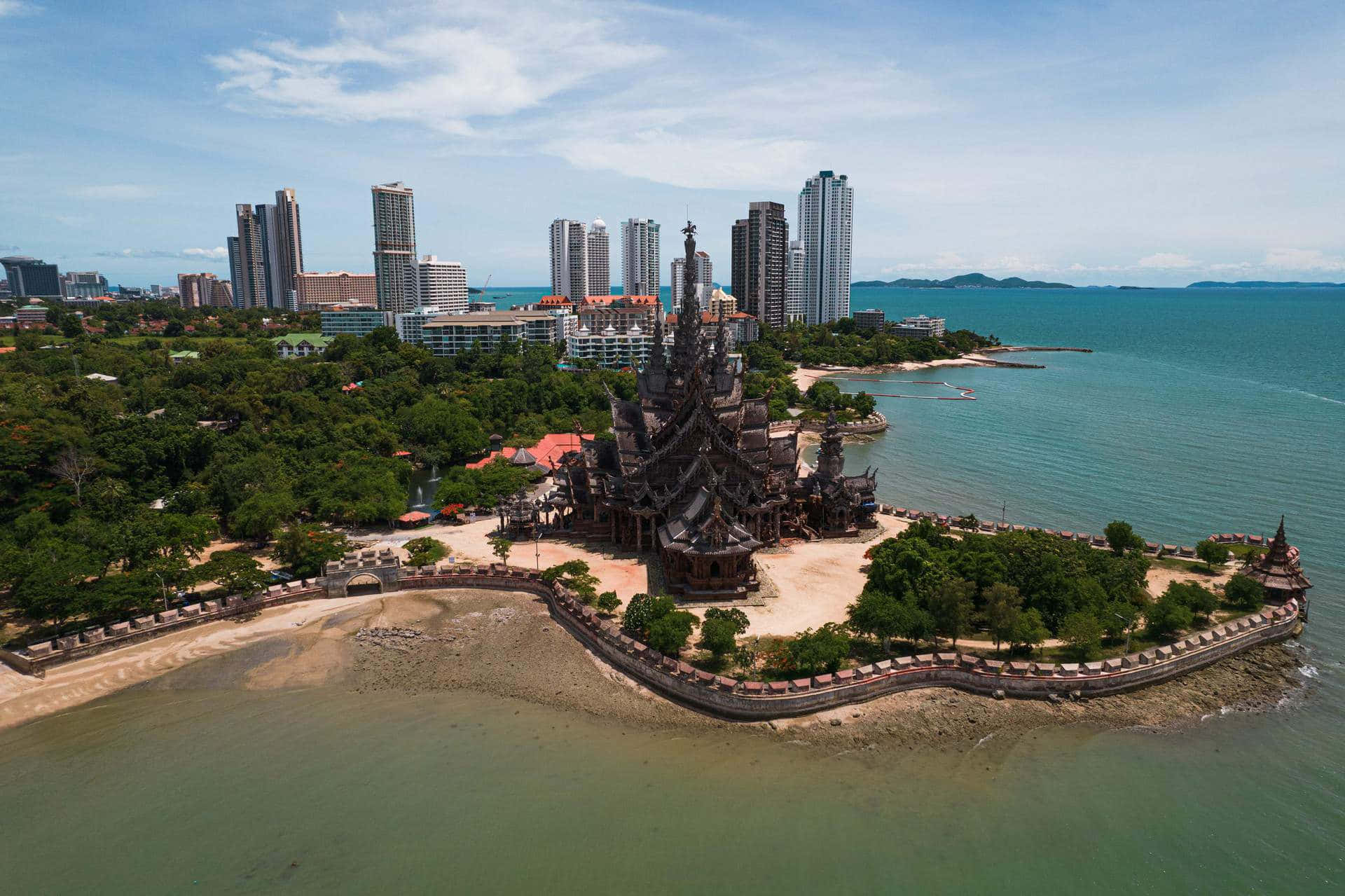 海灘 桌布 壁紙 — 海岸半島上深色木造真理寺廟的鳥瞰圖，背景有現代摩天大樓和碧綠海水。