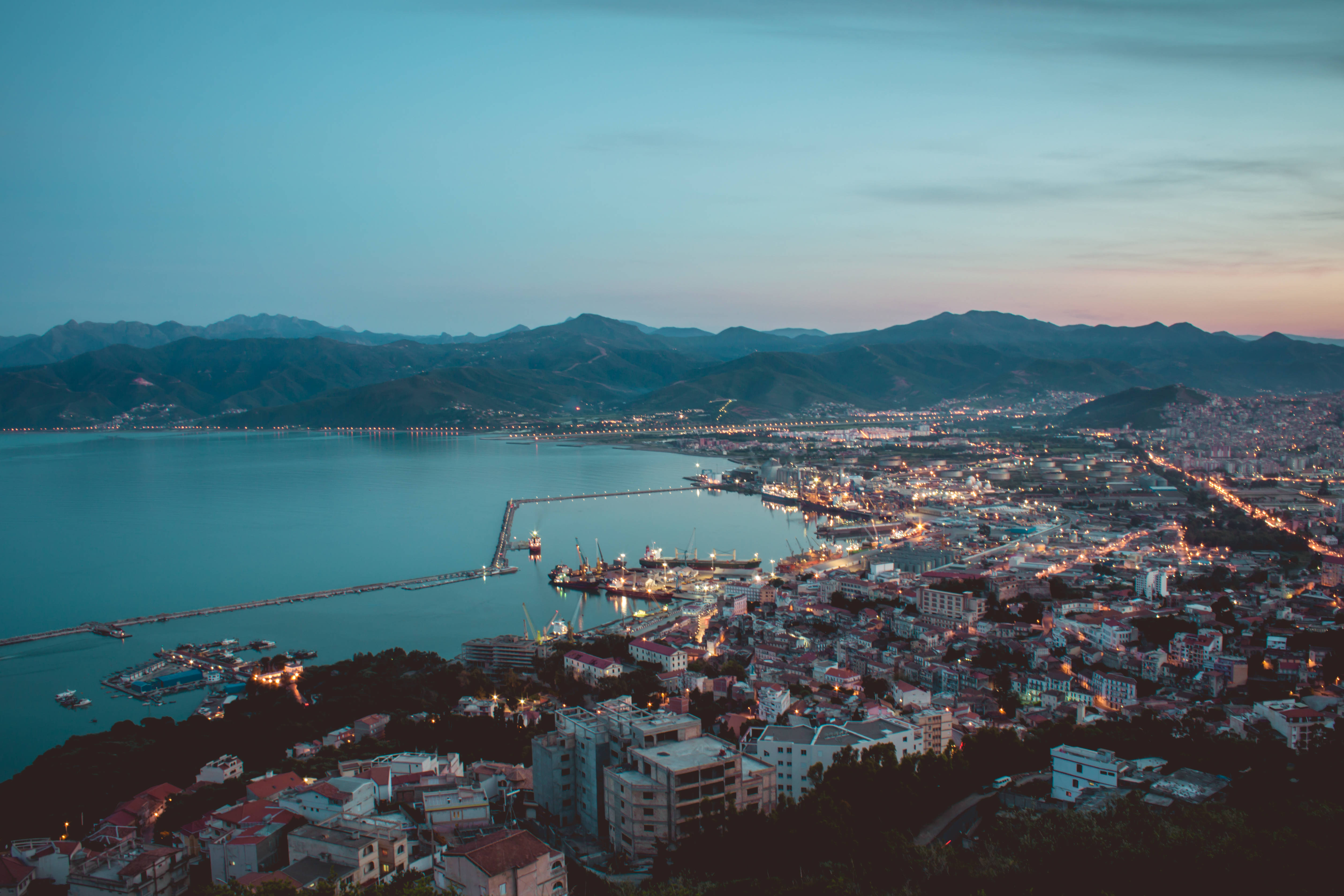 Algeria 桌布 壁紙 — 海岸城市在黃昏時亮起燈光，山巒在平靜的海面後方隱現，天空柔和暮色。