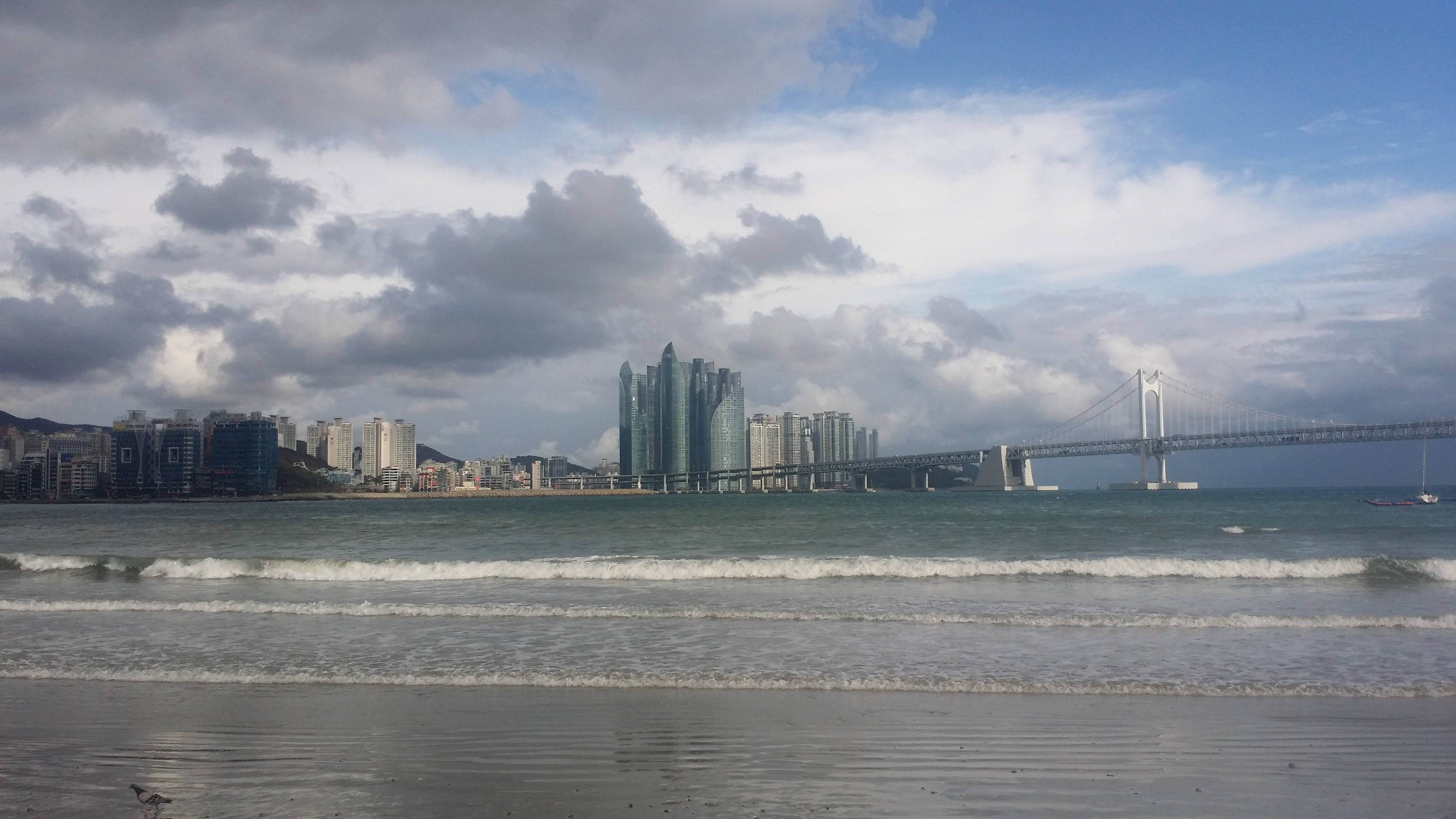 Busan 桌布 壁紙 — 海岸城市景觀，現代摩天大樓和懸索橋，天空多雲，海浪拍打沙灘。