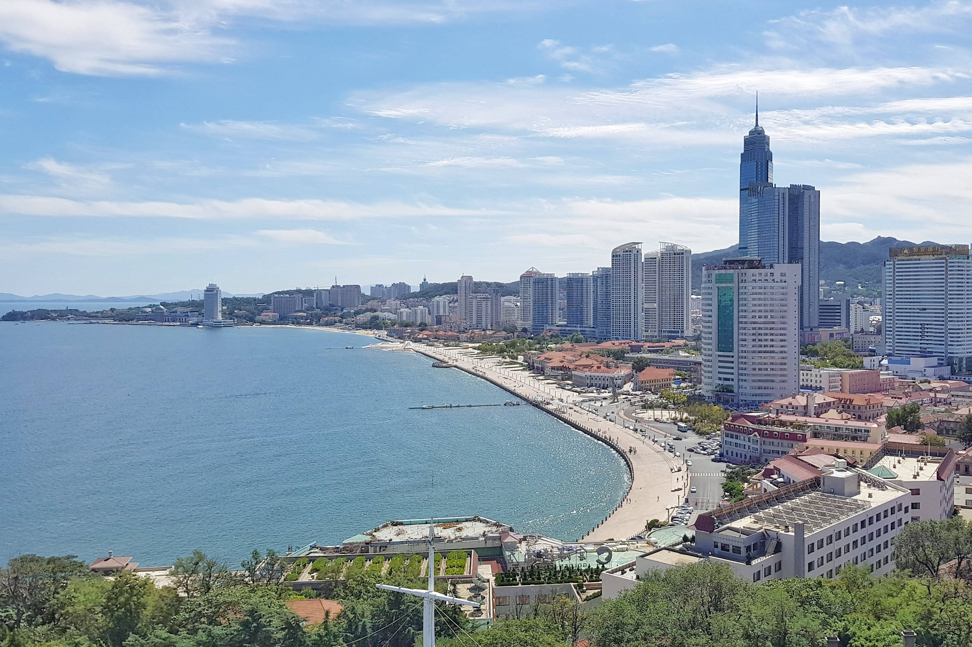 中國 桌布 壁紙 — 海岸城市的全景視圖，現代摩天大樓沿著彎曲的海灘排列，在明亮的藍天白雲下。