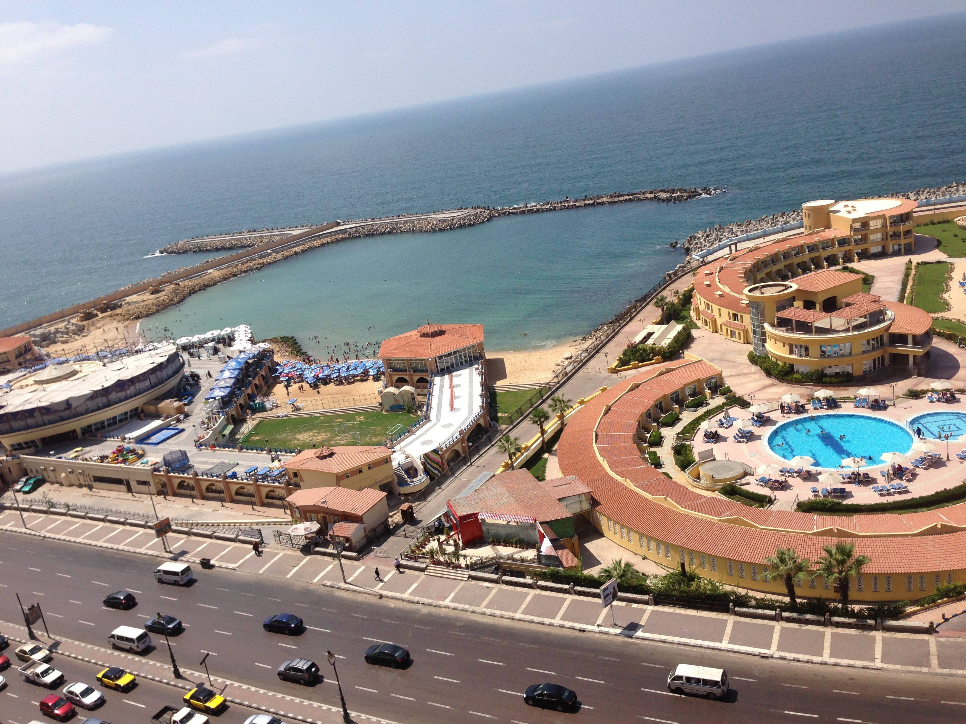 Alexandria 桌布 壁紙 — 海岸度假村的鳥瞰圖，有彎曲的黃色建築物、游泳池和一個延伸到碧綠海洋的碼頭，在晴朗的藍天下。