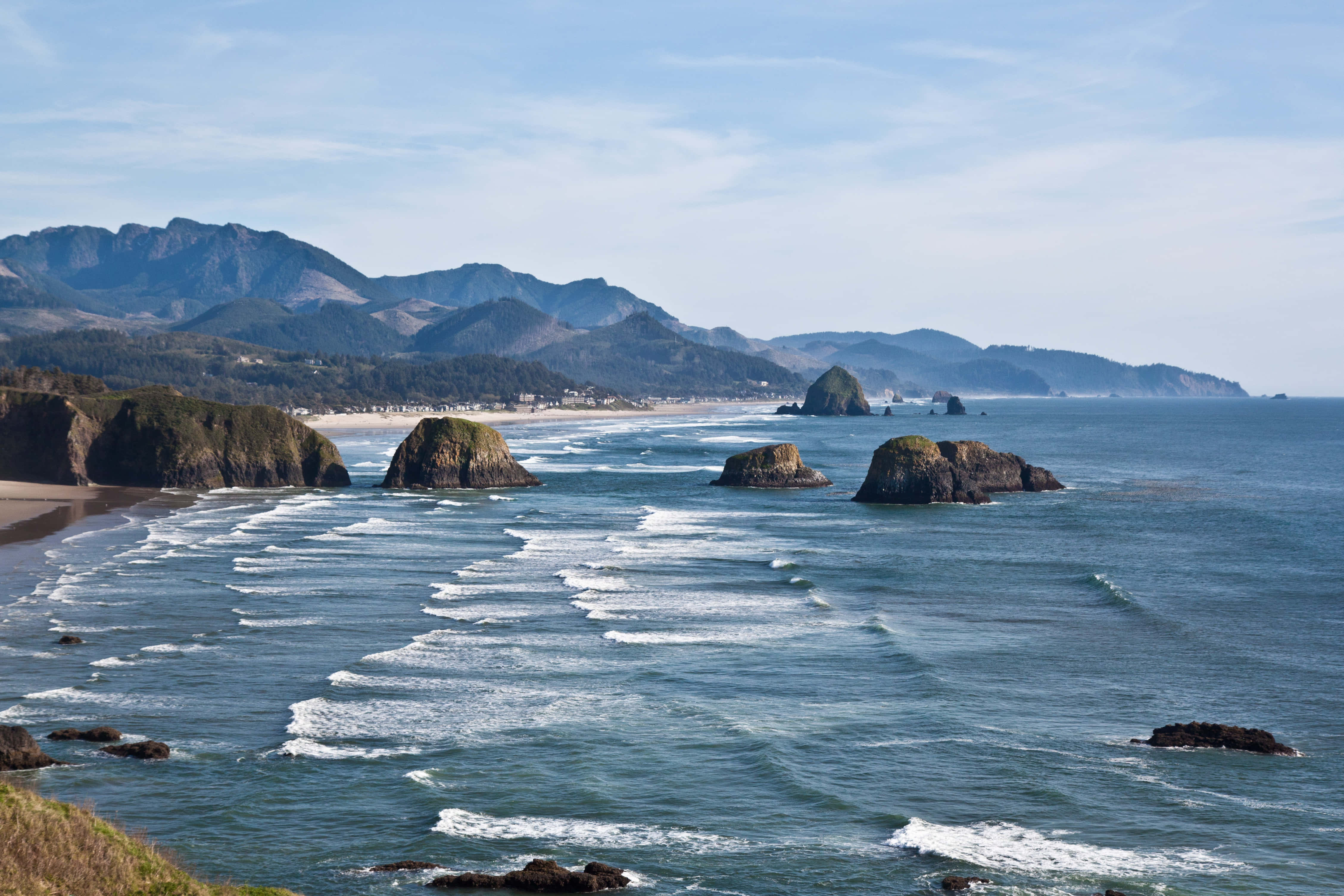 Oregon Coast 桌布 壁紙 — 海岸景色，海浪拍打岩石露頭和遠處山脈，天空清澈湛藍。