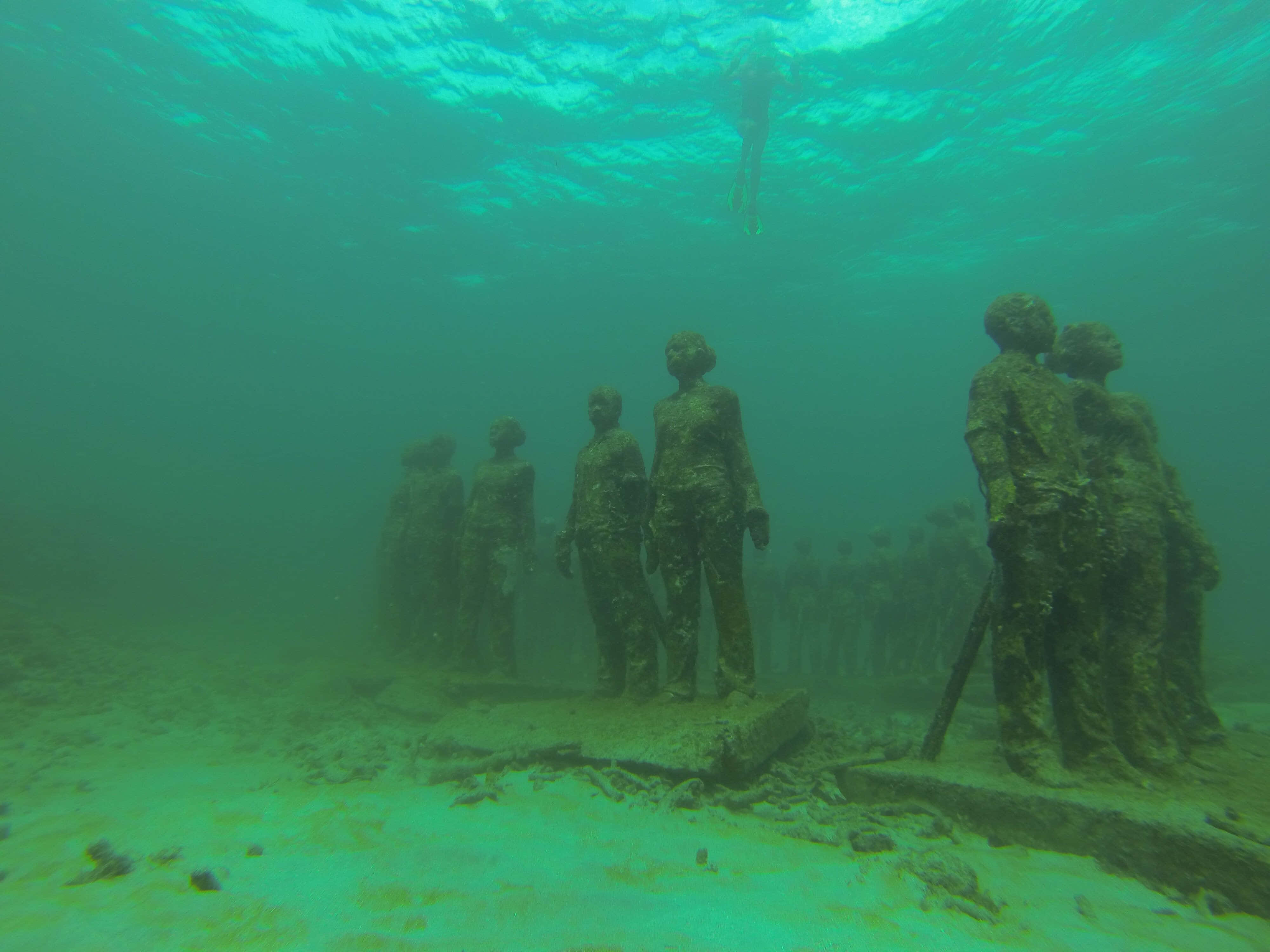 Scary Underwater 桌布 壁紙 — 海底的人像雕像莊嚴地矗立在沙質海床上，一名潛水員在碧綠色的海水中從上方滑過。