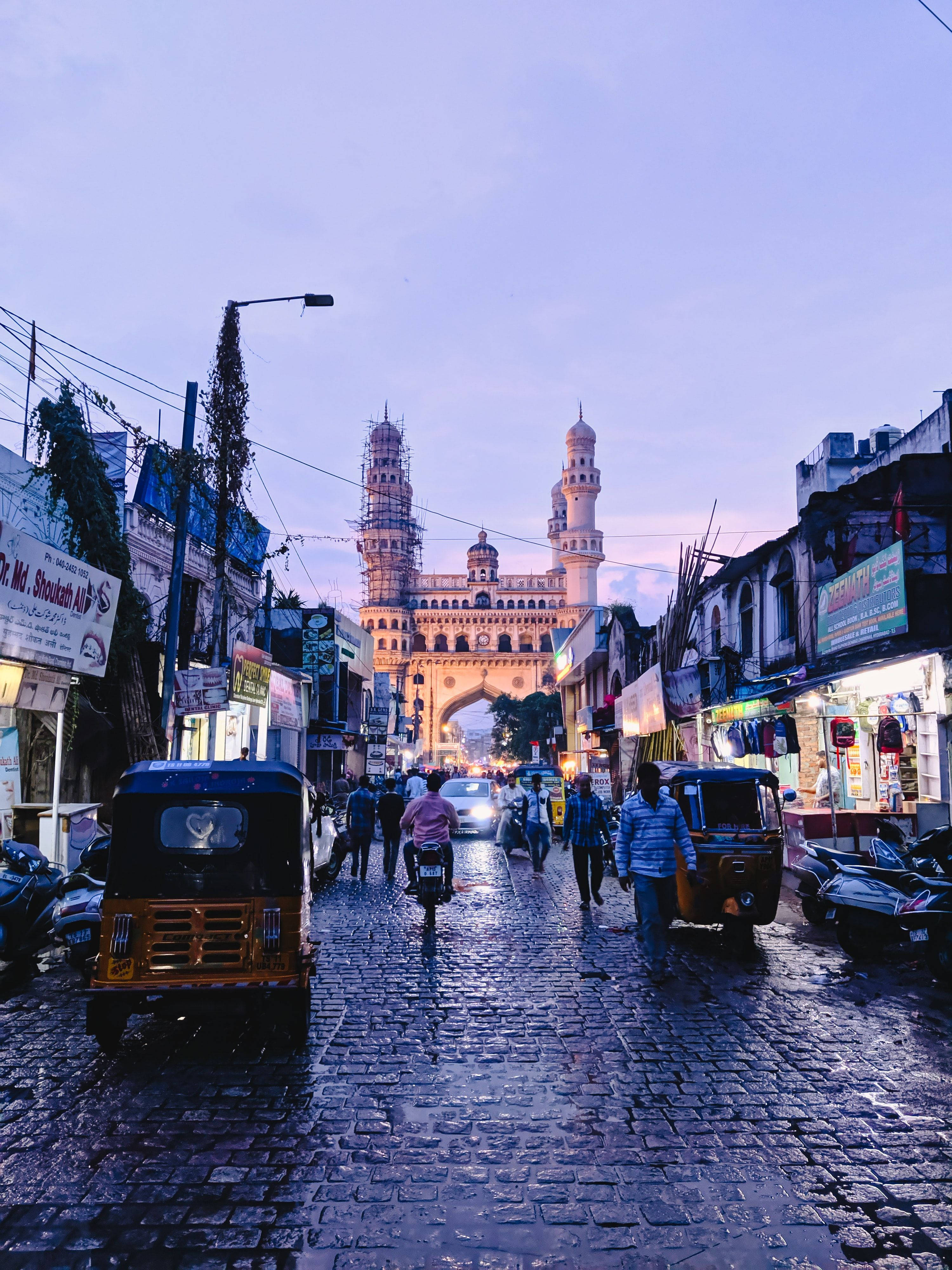 Hyderabad 桌布 壁紙 — 海德拉巴一條濕漉漉的鵝卵石街道在黃昏時分通向燈火通明的查爾米納爾，有自動三輪車和行人。