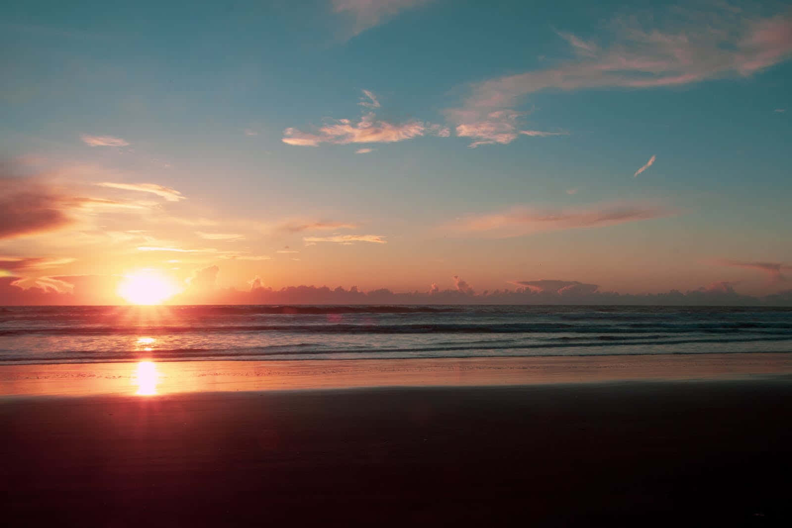 Cute Sunset 桌布 壁紙 — 海洋上寧靜的日落，金色光線反射在濕沙和柔和的粉橙色雲朵上。